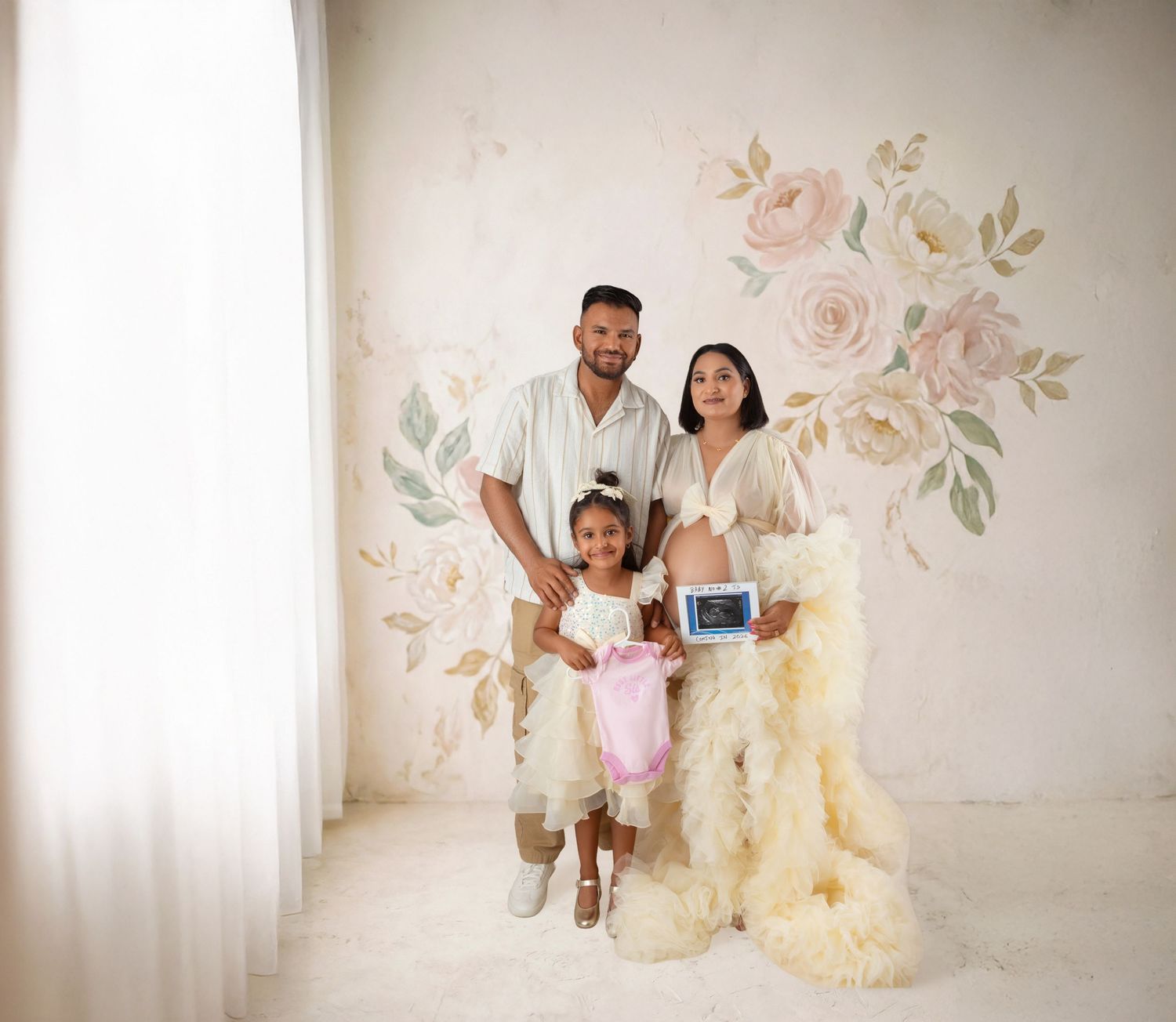Family maternity photo with parents and young daughter posing in front of floral wall mural, mother in elegant gown.