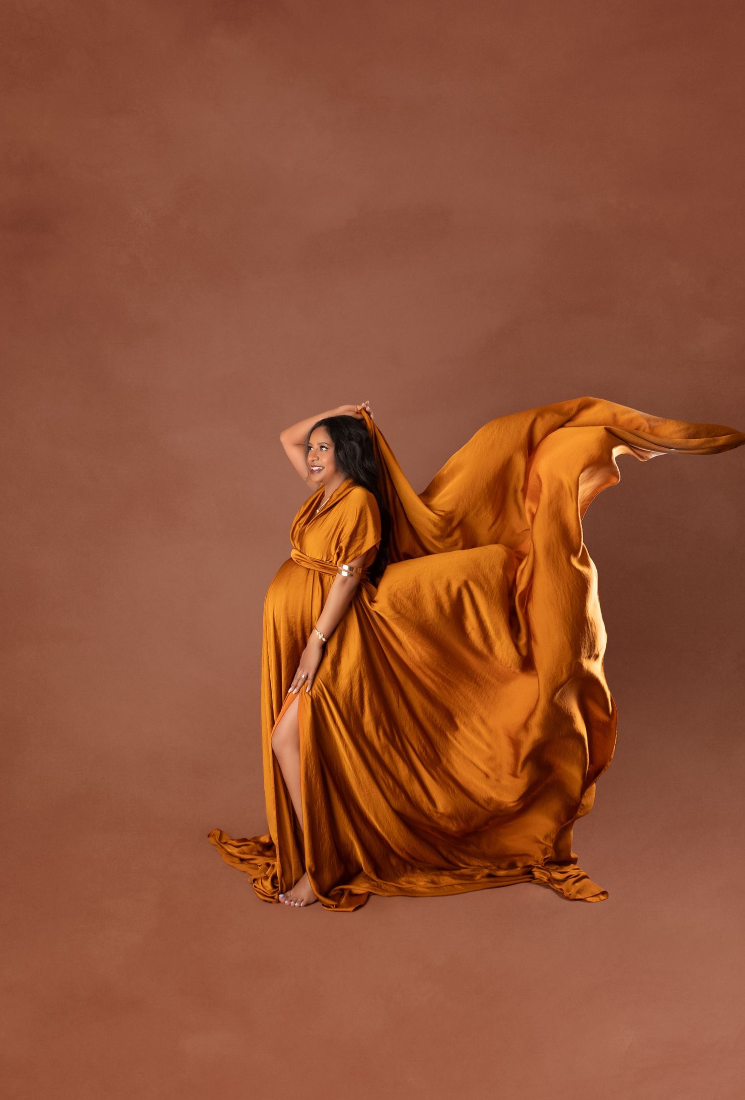Pregnant woman in flowing burnt orange maternity gown with dramatic fabric movement against warm brown studio backdrop.