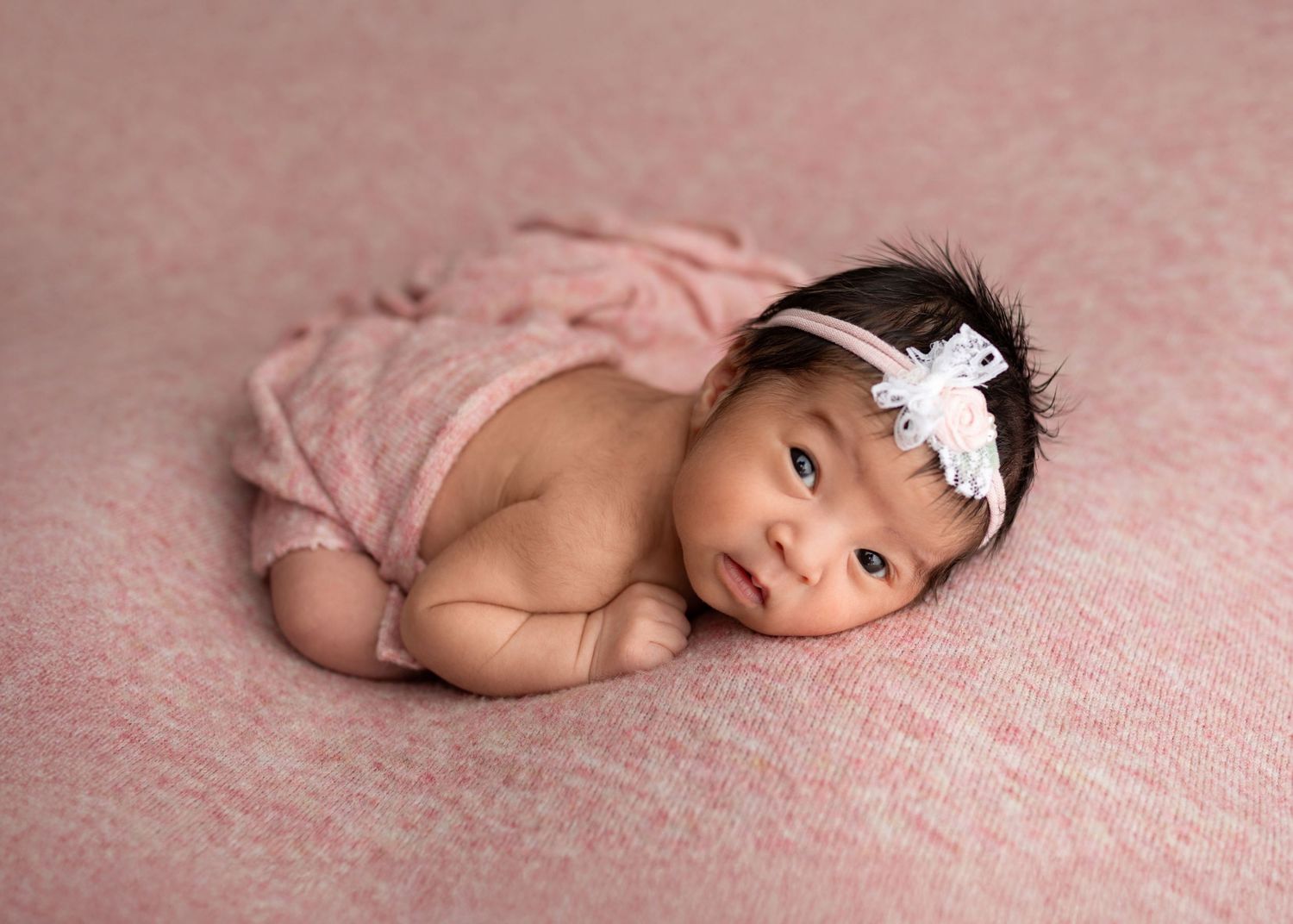 Newborn baby girl in pink wrap lying on pink blanket with white floral headband.