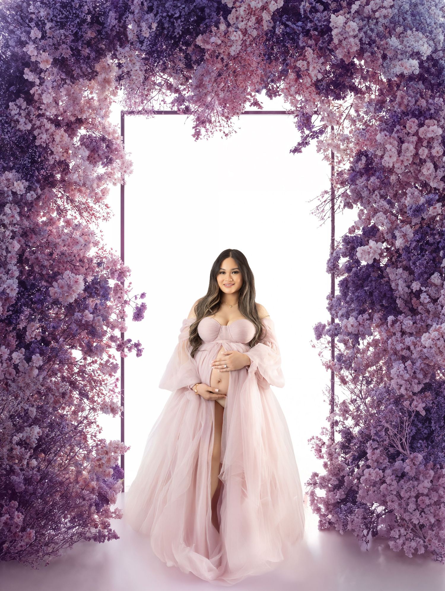 Pregnant woman in pink tulle gown poses under purple floral arch for elegant maternity portrait.