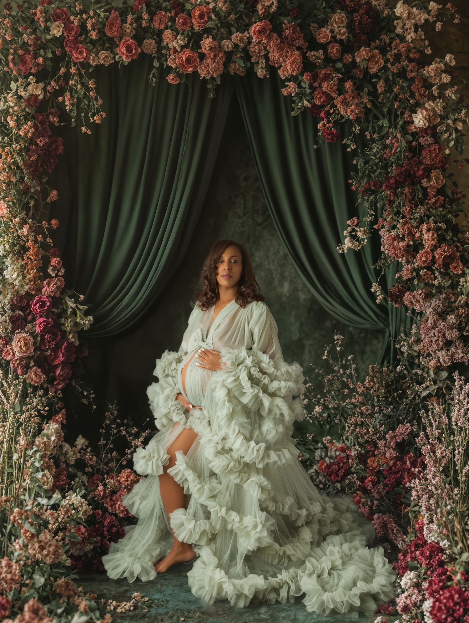 Pregnant woman in white ruffled gown poses in floral arch with green drapes and pink roses backdrop.