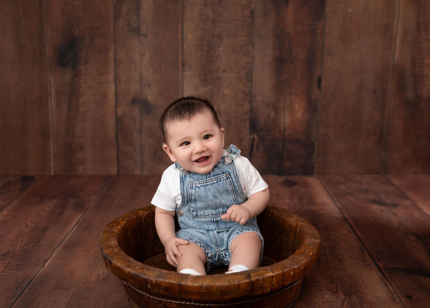 Smiling baby in denim overalls sitting in a wooden bowl on a rustic wood floor backdrop.