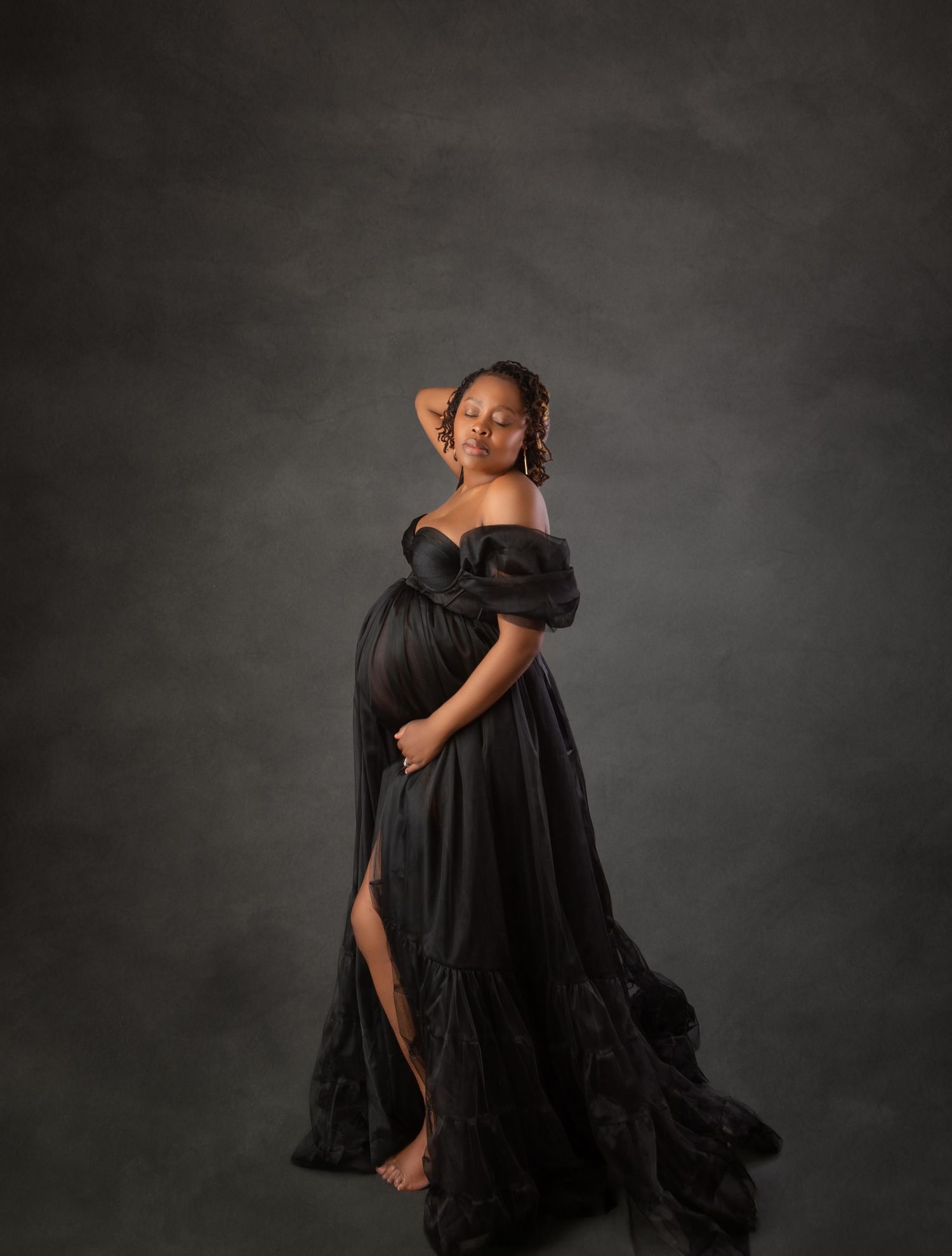 Pregnant woman in black off-shoulder tulle gown with slit poses on gray studio backdrop.