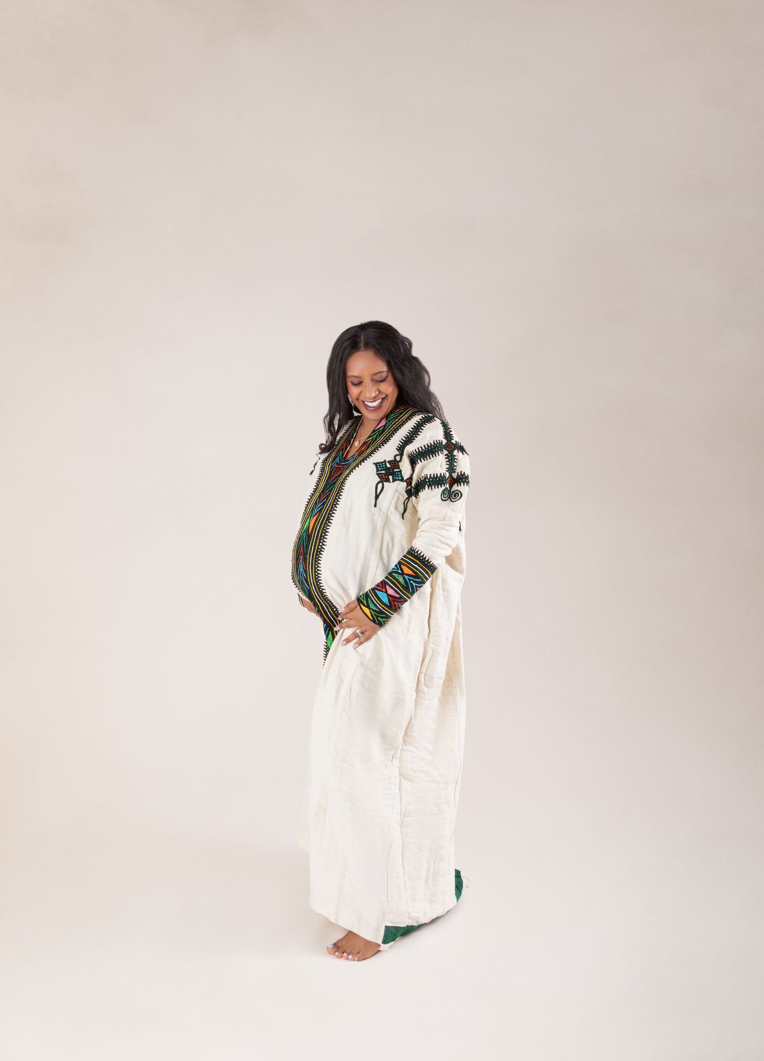 A smiling pregnant woman in traditional white Ethiopian dress with colorful embroidery poses in studio.