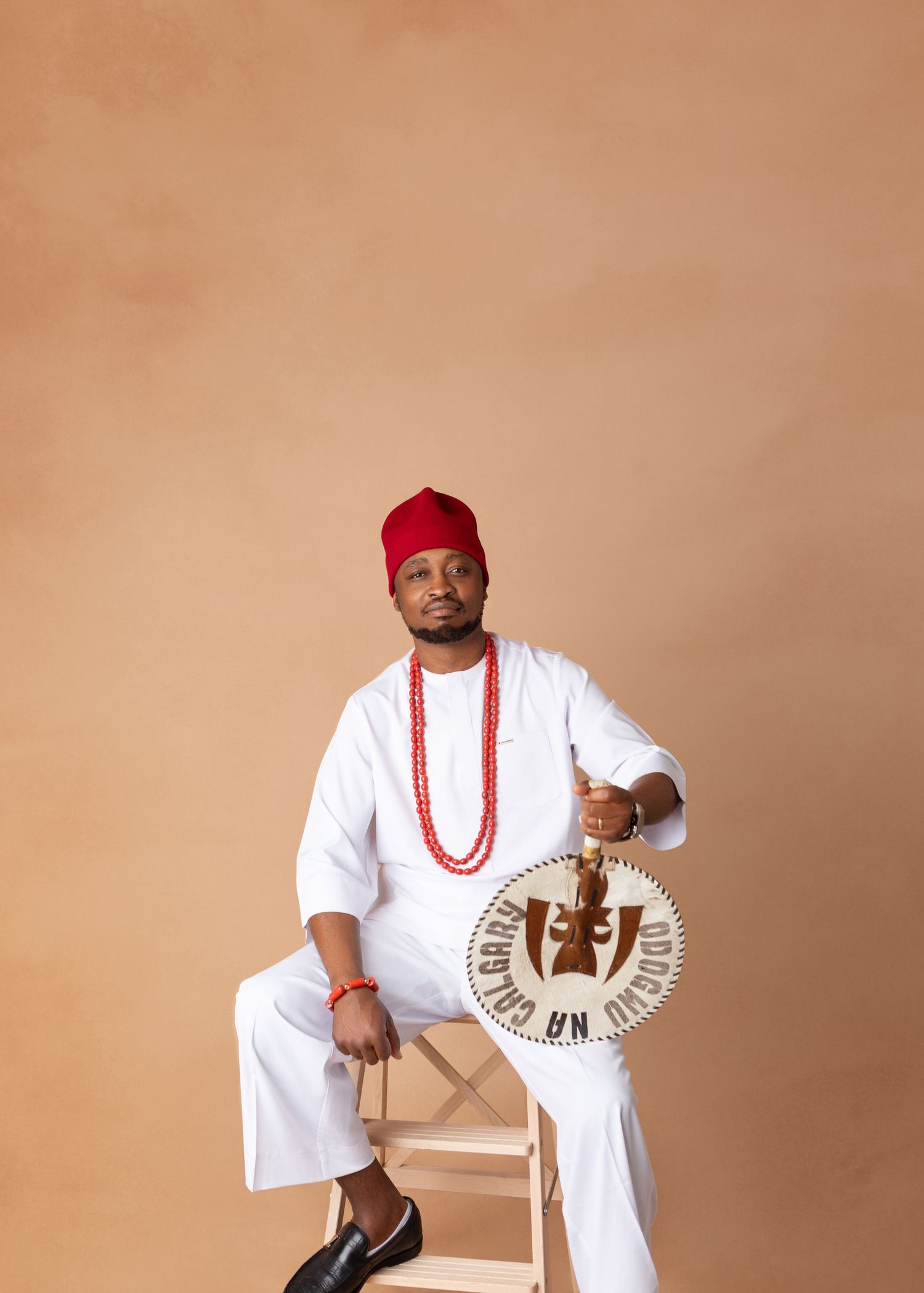 Man in white traditional Nigerian outfit with red cap sitting on chair holding ceremonial drum.
