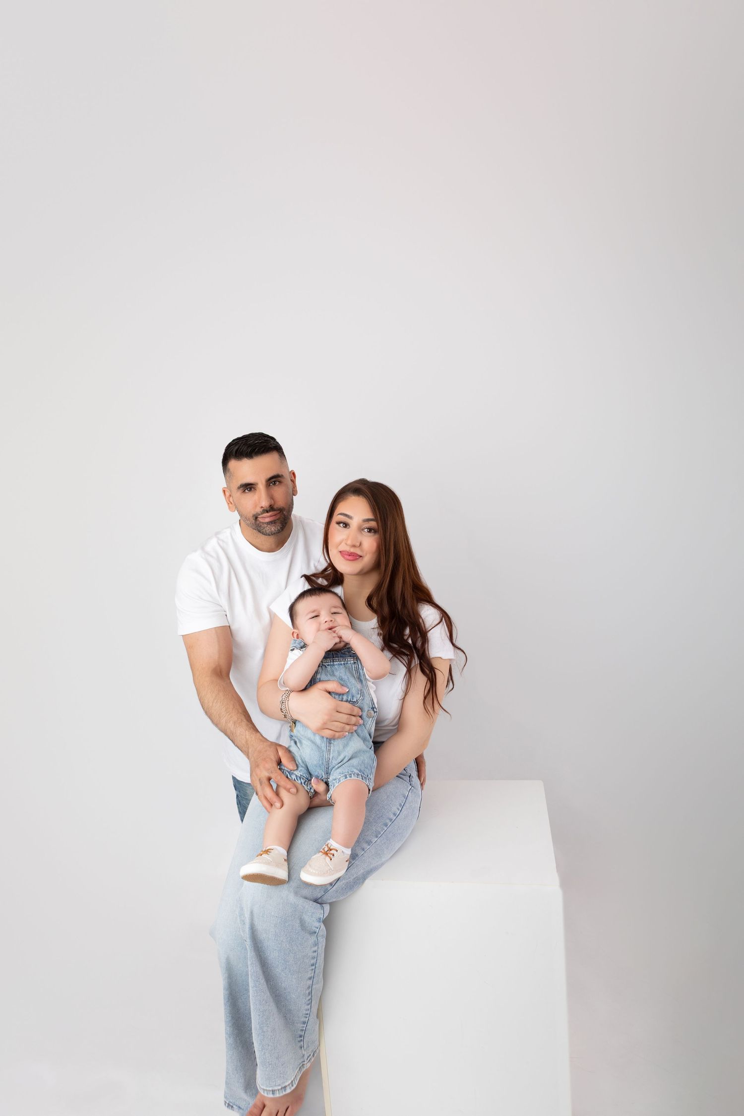 Family of three with parents in white shirts sitting with baby on white cube in studio.