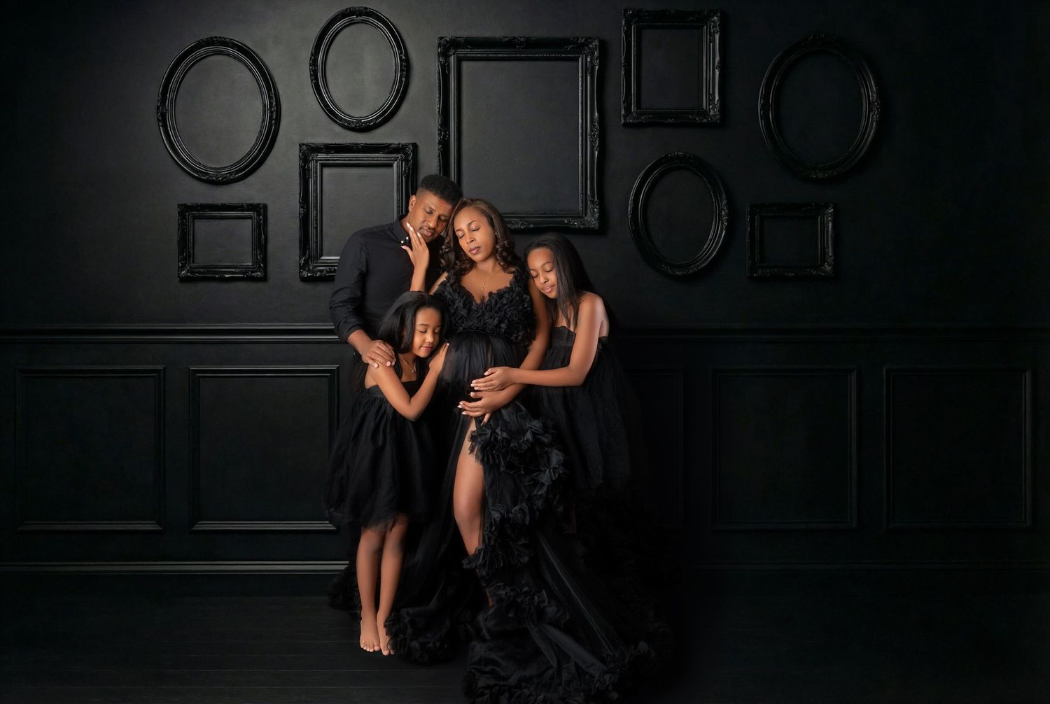 Family of four in black attire poses together against dark wall for maternity portrait.