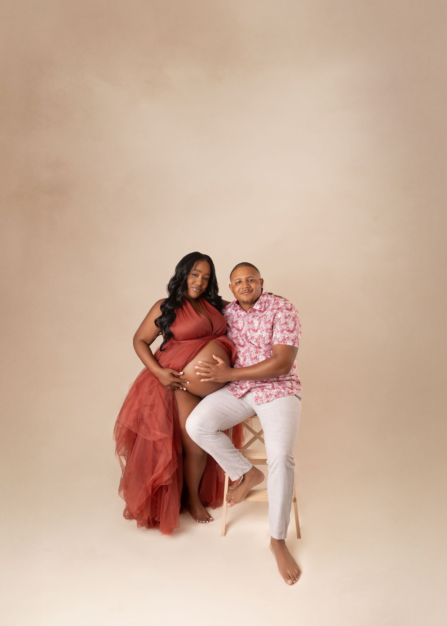 Expecting couple poses together on stool against warm beige backdrop in maternity studio photo.