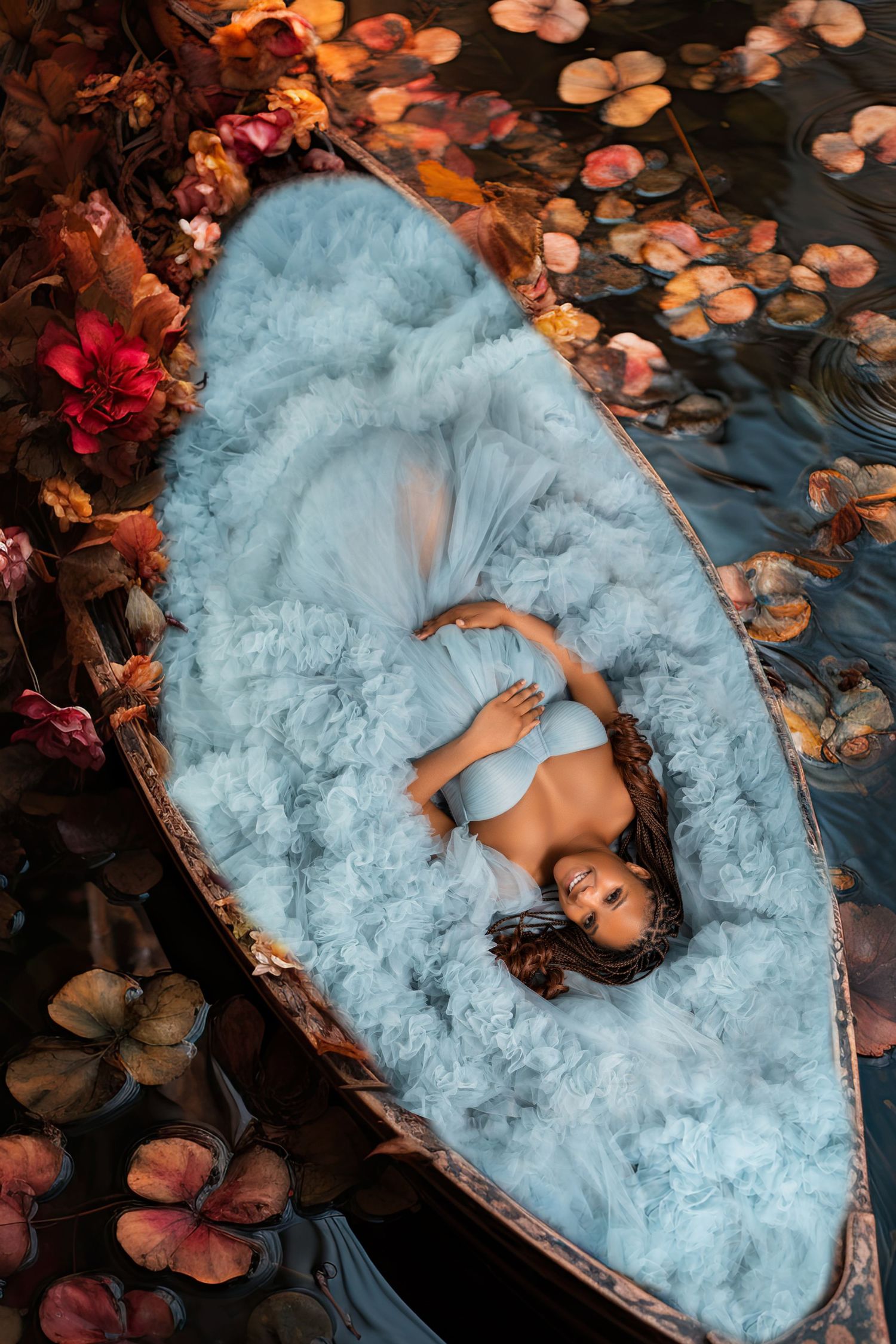Woman in blue tulle gown lying in a flower-adorned boat surrounded by colorful lily pads on dark water.