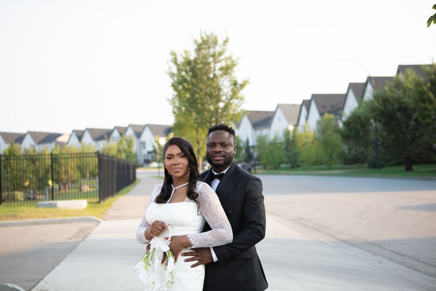 Bride and groom posing outdoors in wedding attire, white gown and black tuxedo, with suburban homes in background.