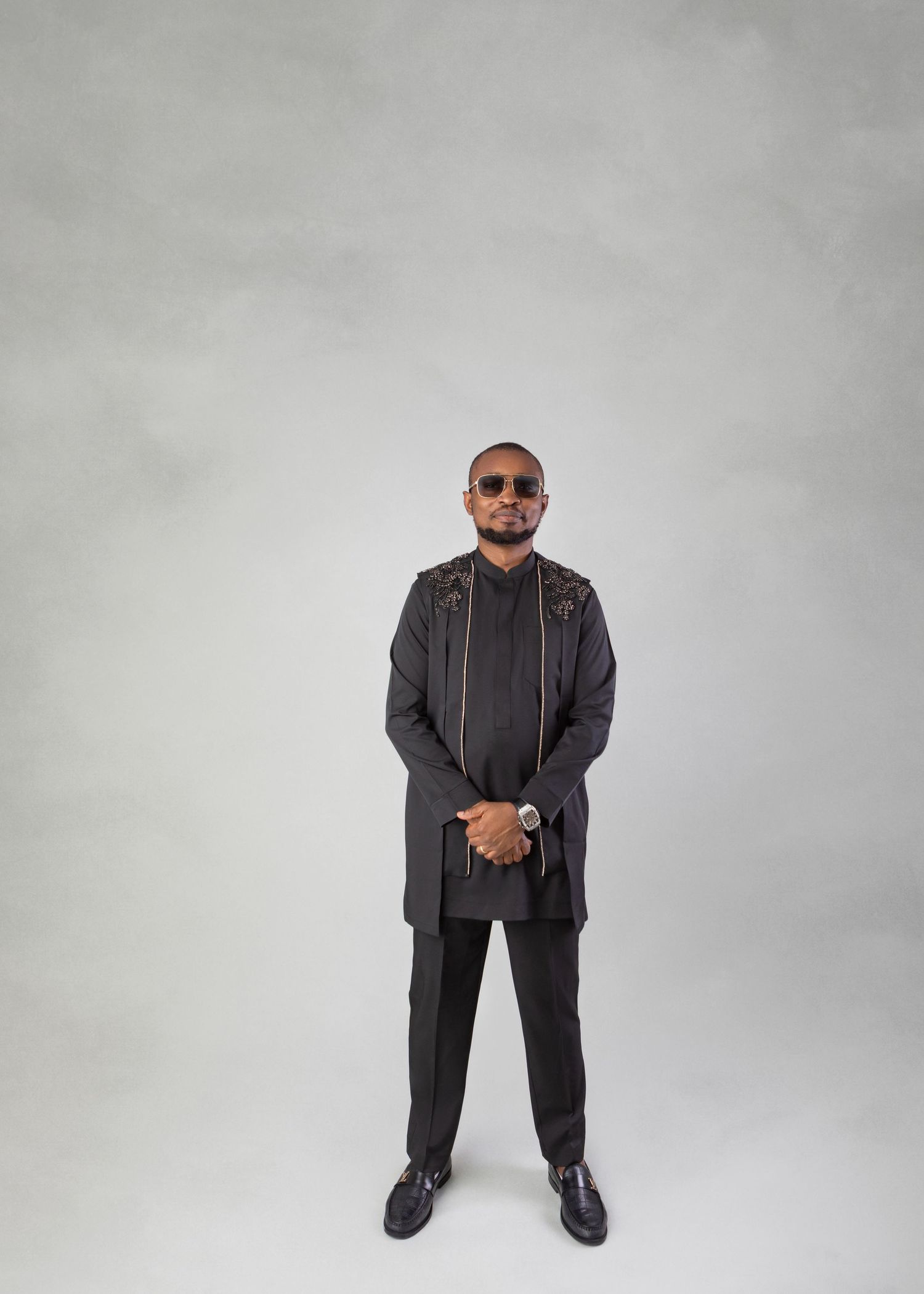 Man in black traditional African attire and sunglasses poses confidently against a neutral gray studio background.