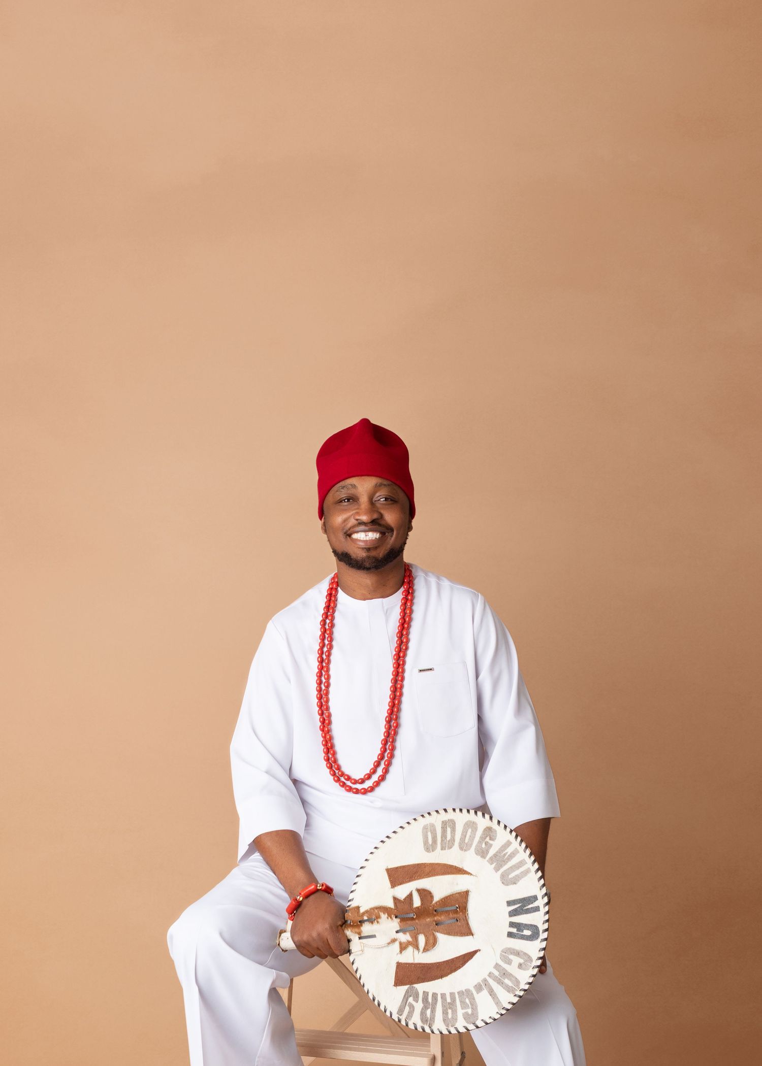 Smiling man in white traditional Nigerian attire with red beaded necklace and cap, holding decorative fan while seated.