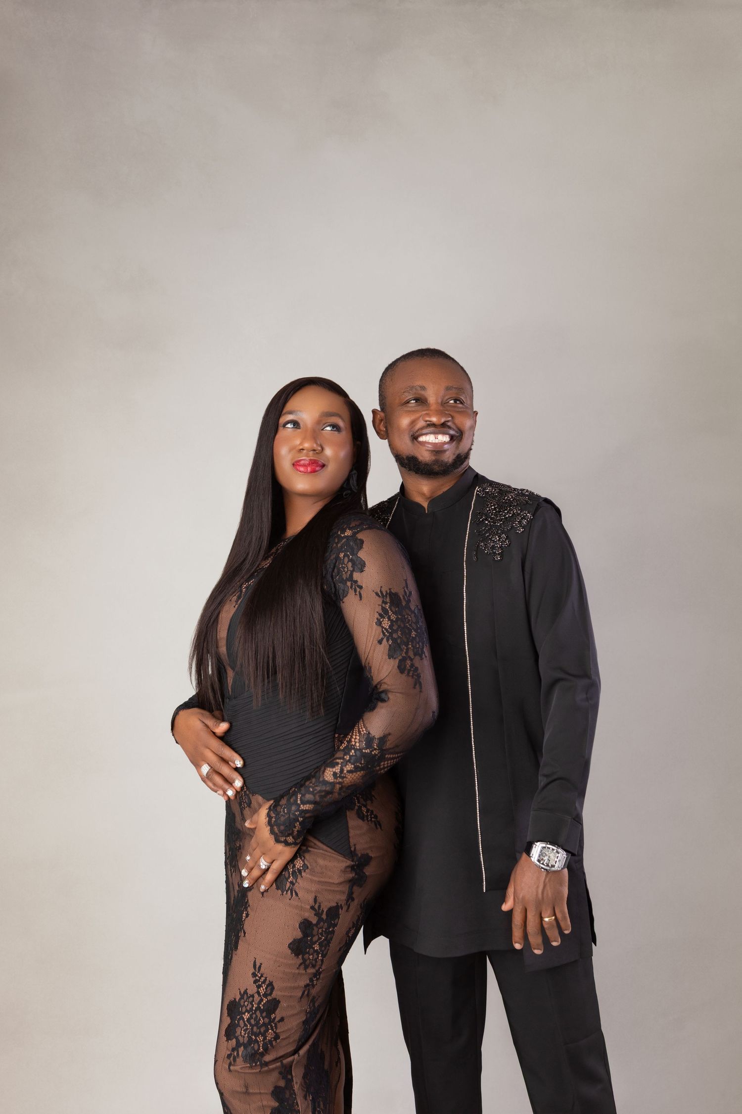 Elegant couple in matching black outfits posing together against a gray studio background.