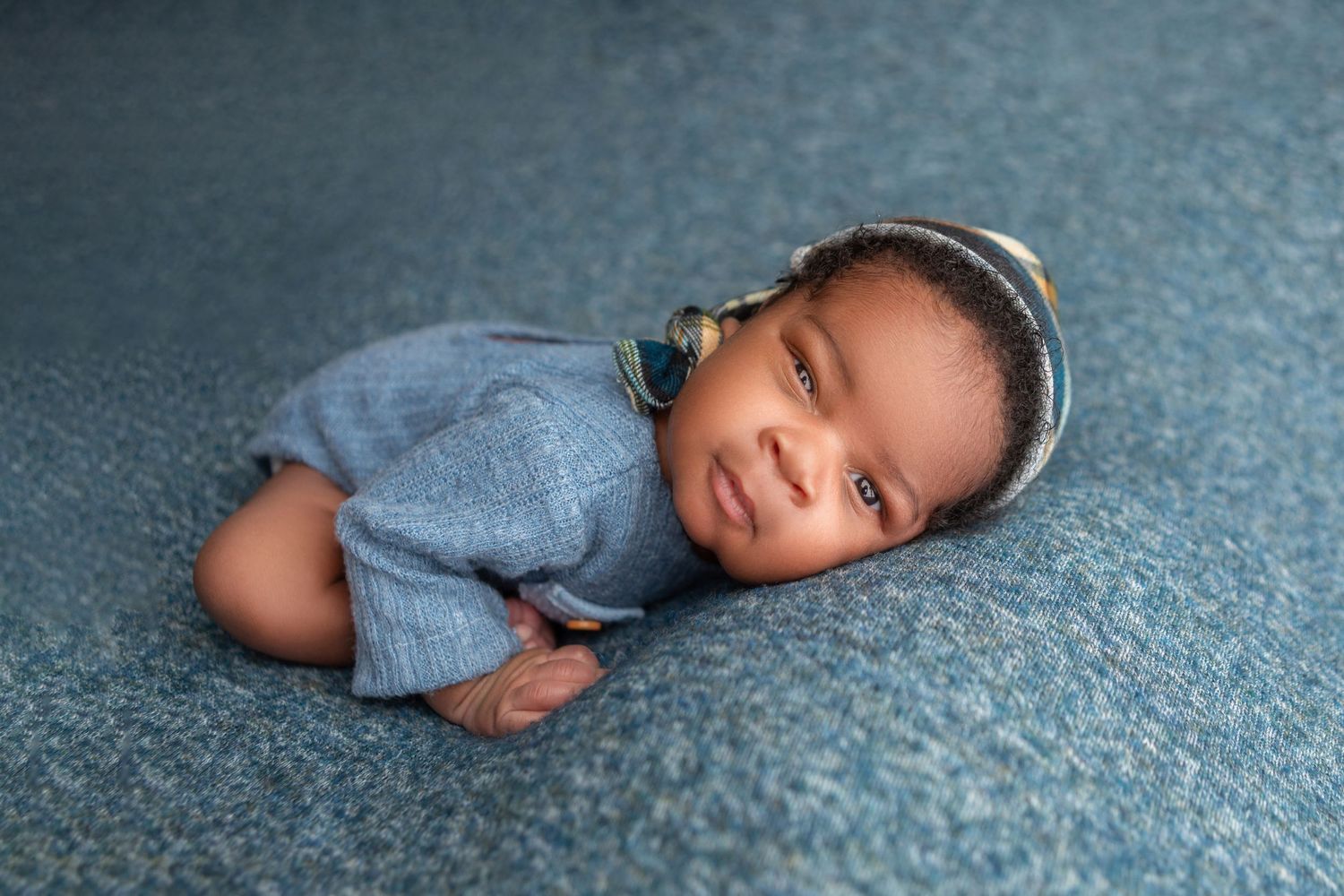 Smiling newborn baby in blue romper and knit hat lying on blue textured backdrop.