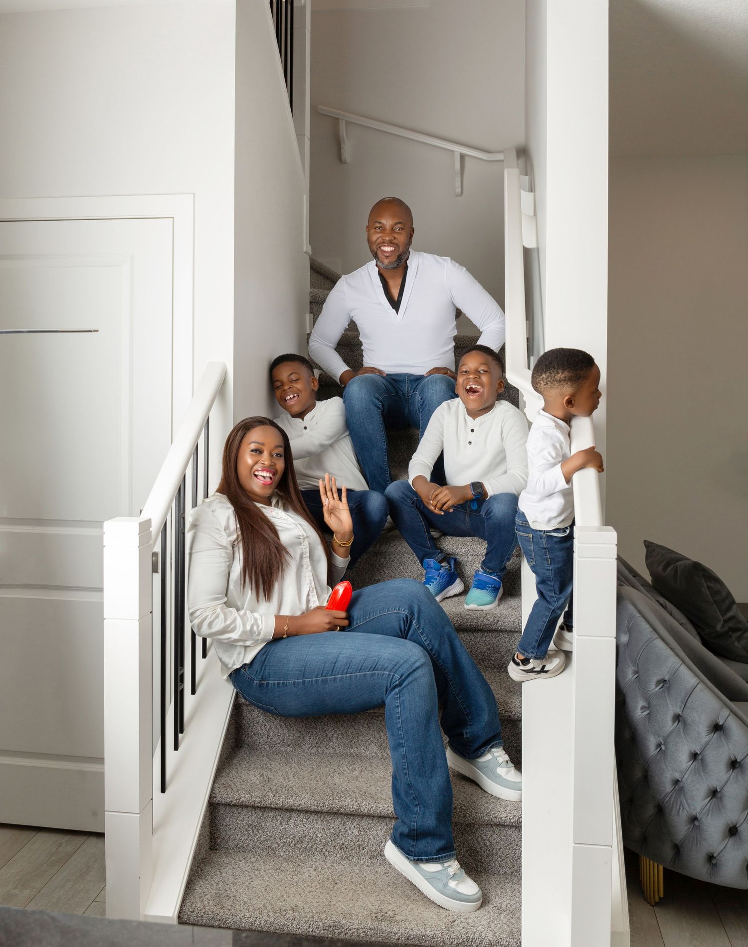 Happy Black family of five posing together on a staircase, wearing jeans and white tops, smiling and waving at the camera.