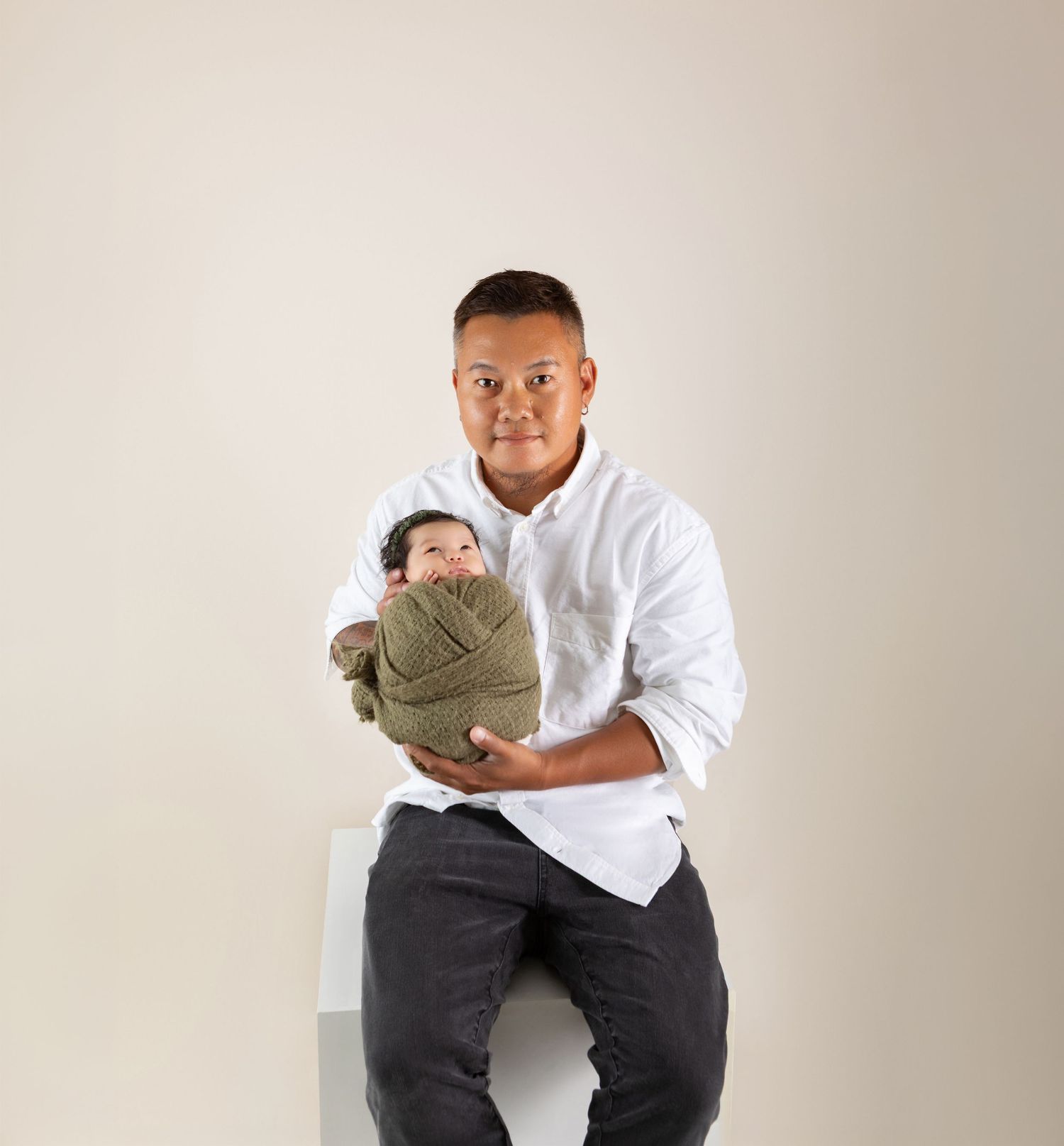 Father in white shirt holds swaddled newborn baby in olive wrap during professional studio portrait.