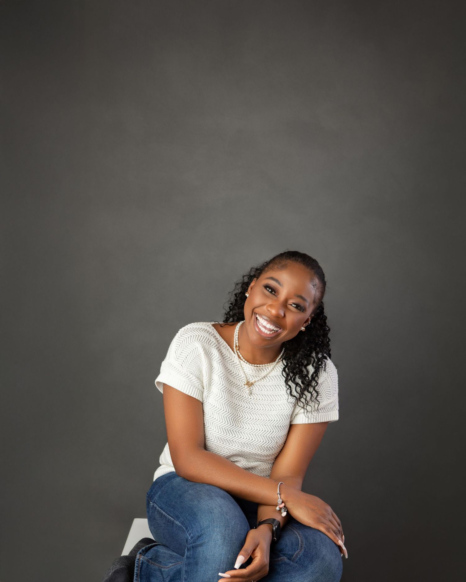 A young woman with curly hair smiles brightly, sitting casually in jeans and a white top against a gray backdrop.