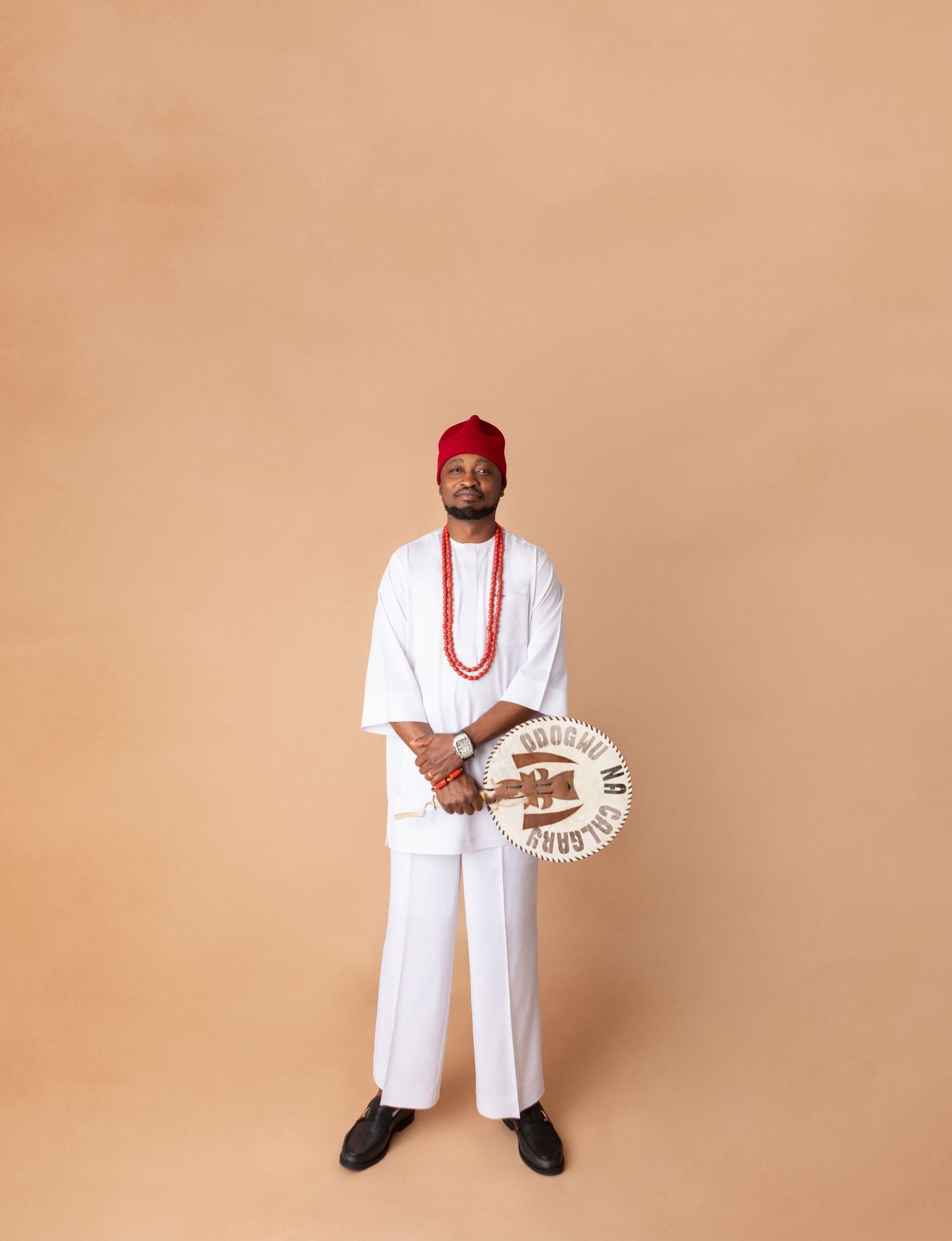 Man in white traditional Nigerian attire with red beaded necklace, red cap, holding a decorative plate, standing against beige background.