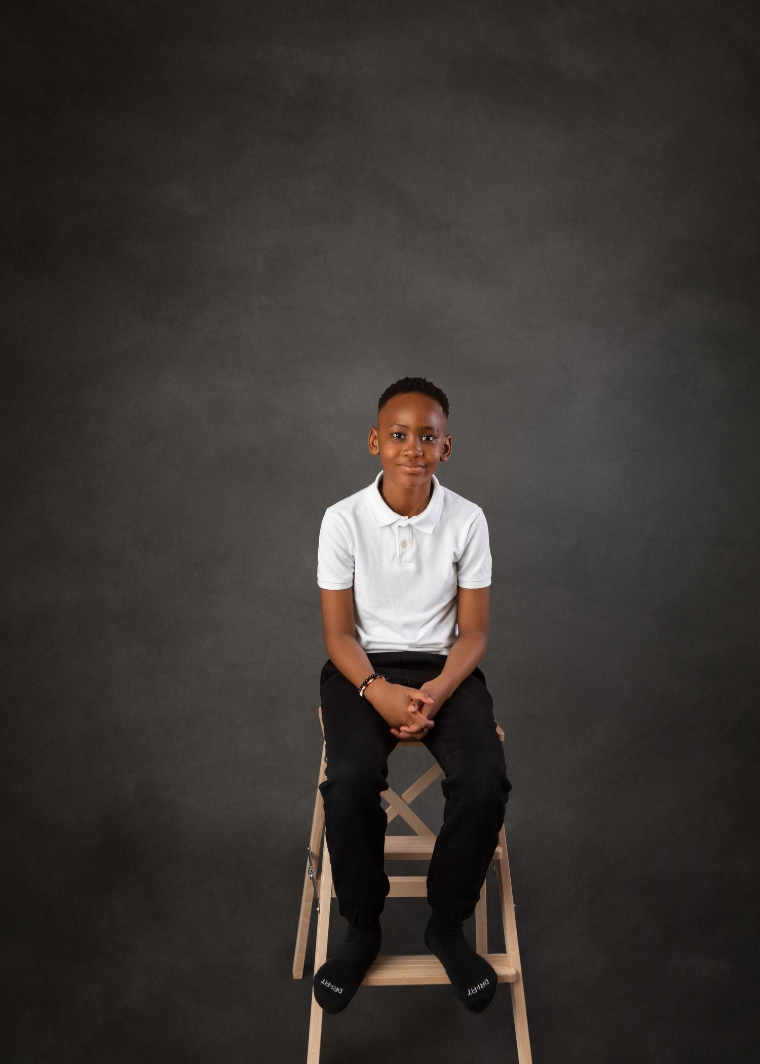 Young person in white polo shirt and black pants sits on a wooden ladder stool against a gray studio backdrop.