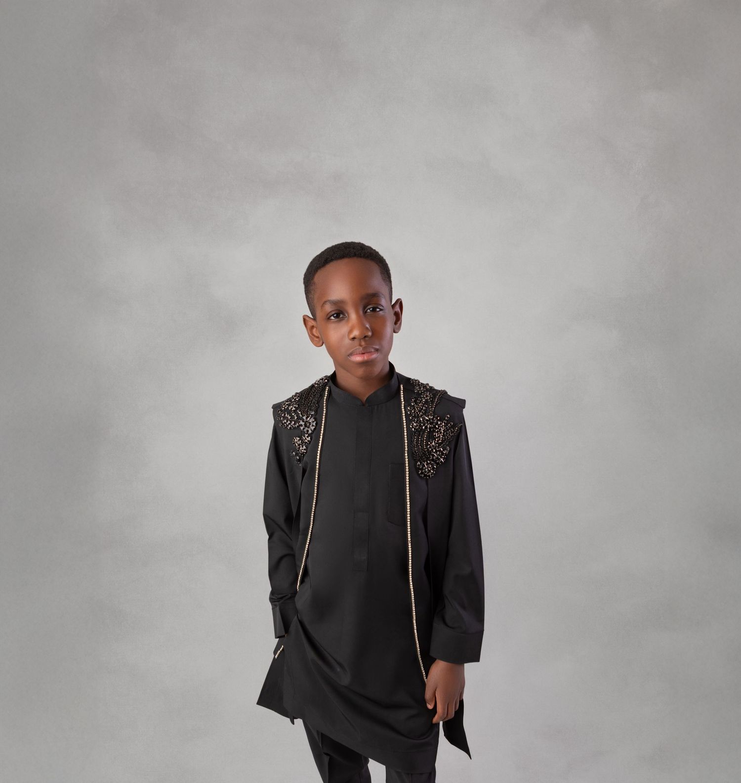 Young boy wearing a black embroidered traditional outfit with gold trim, posing against a gray backdrop.