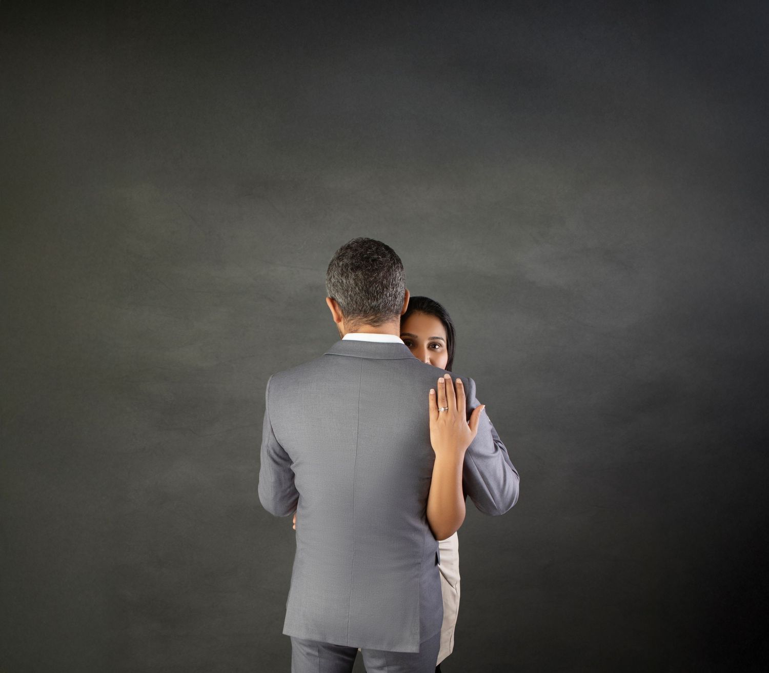 A woman peeks around a man in a gray suit against a dark background, placing her hand on his back.