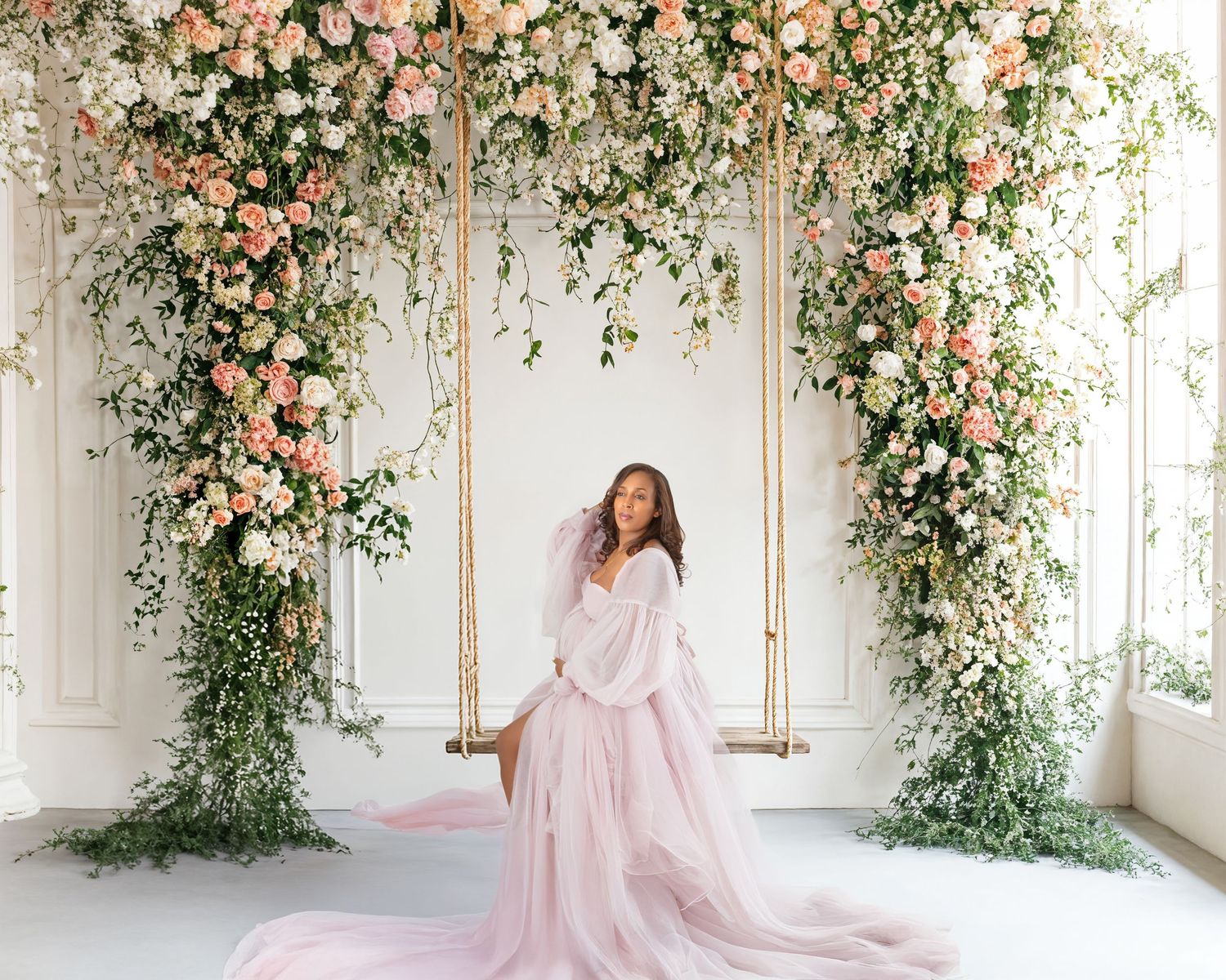 Woman in pink gown sits on floral swing surrounded by lush roses and greenery in elegant white room.
