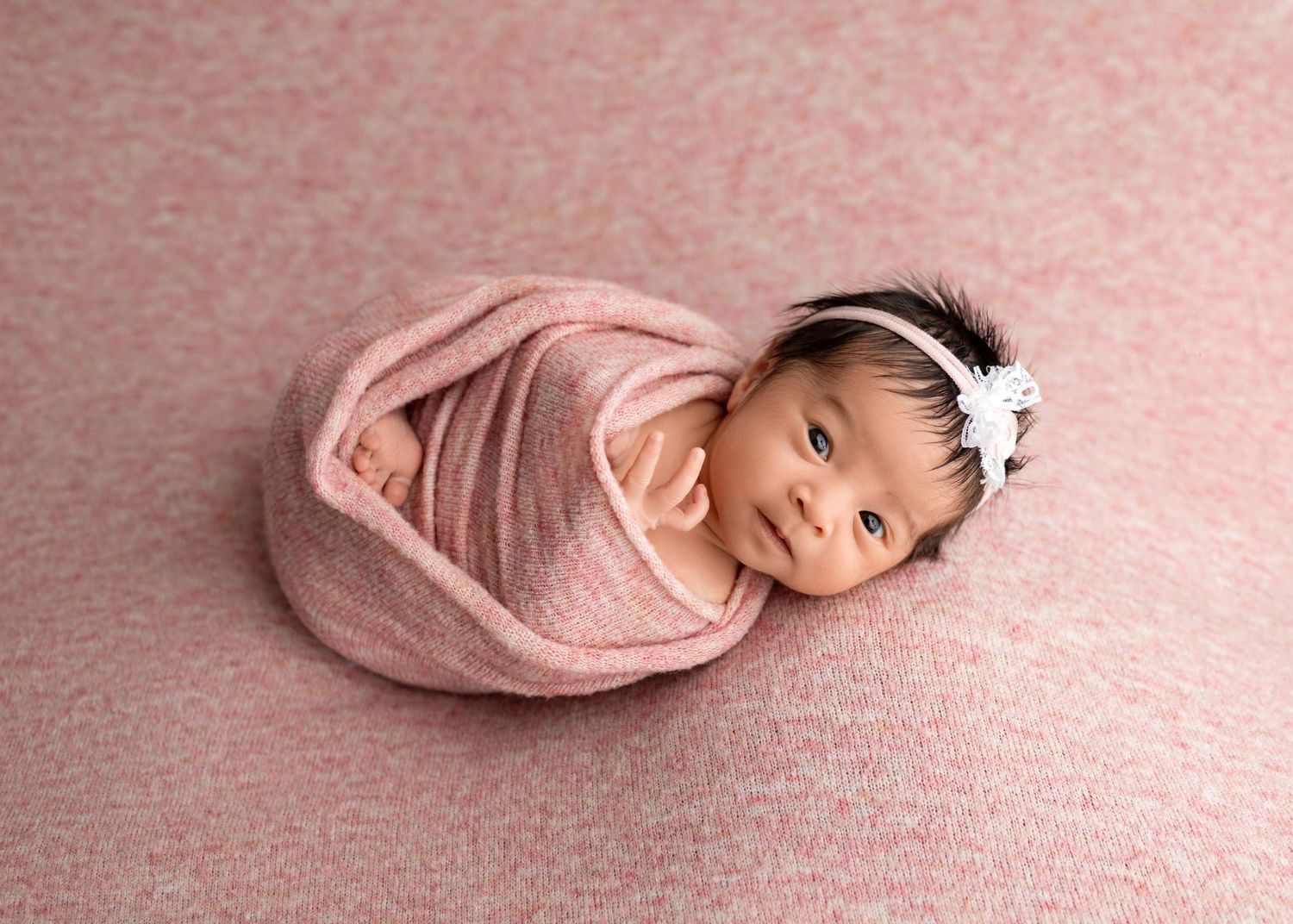 Smiling newborn baby swaddled in pink wrap with white flower headband on pink backdrop.