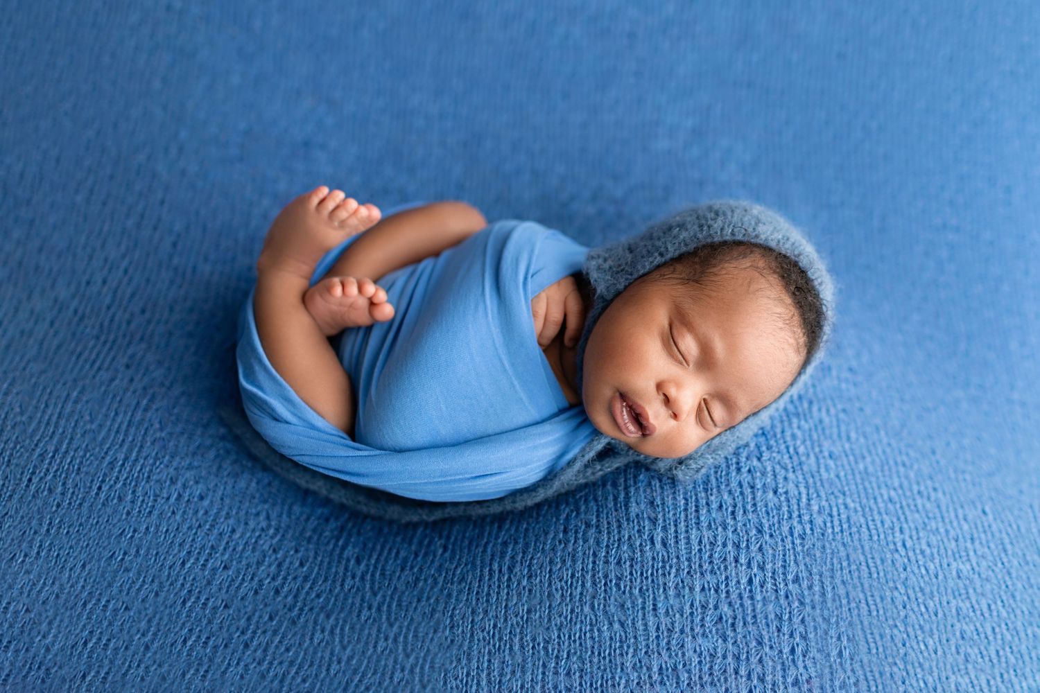 Sleeping newborn baby swaddled in blue wrap with knit bonnet on blue textured background.