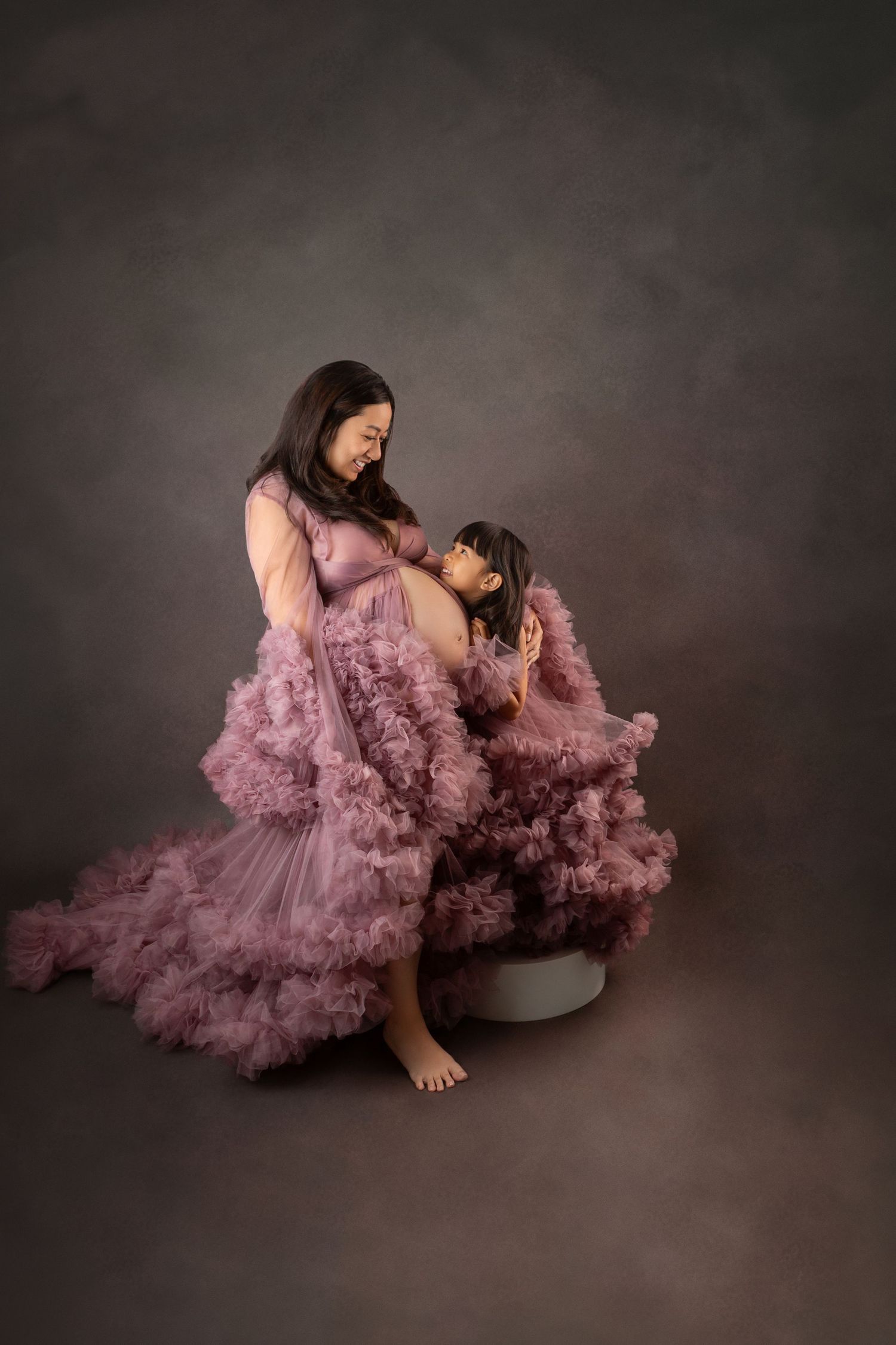 Pregnant Asian woman and daughter share tender moment in matching mauve ruffle tulle maternity gowns.