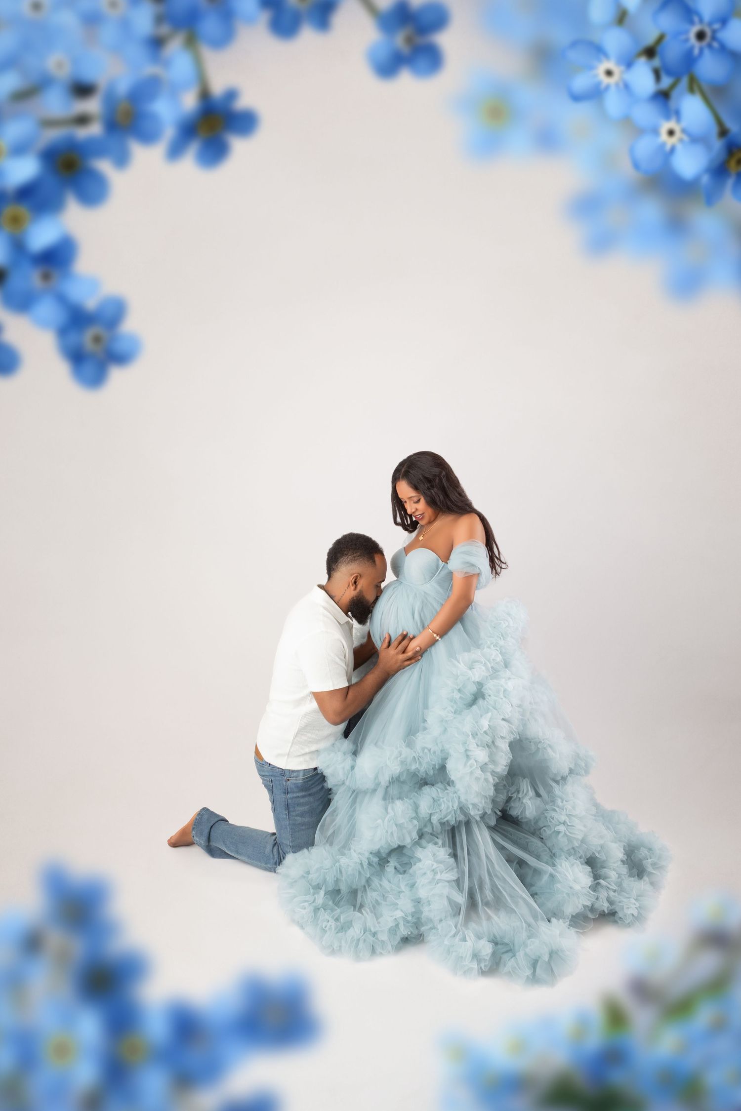 A man kneels kissing a pregnant womans belly as she wears a blue tulle gown with floral frame.