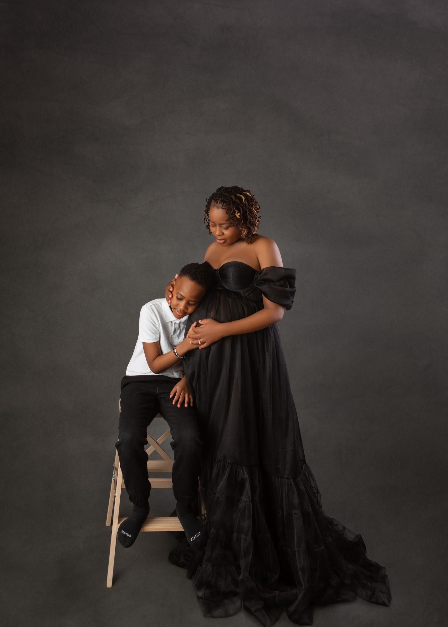 Pregnant woman in black off-shoulder gown poses with young boy on gray studio backdrop.