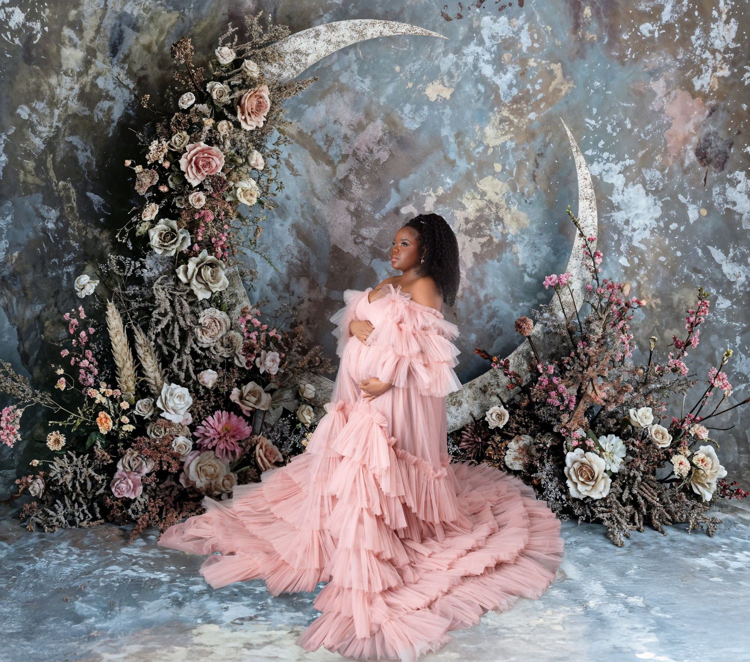 Pregnant woman in blush pink ruffled gown poses by a floral crescent moon on a painted backdrop.