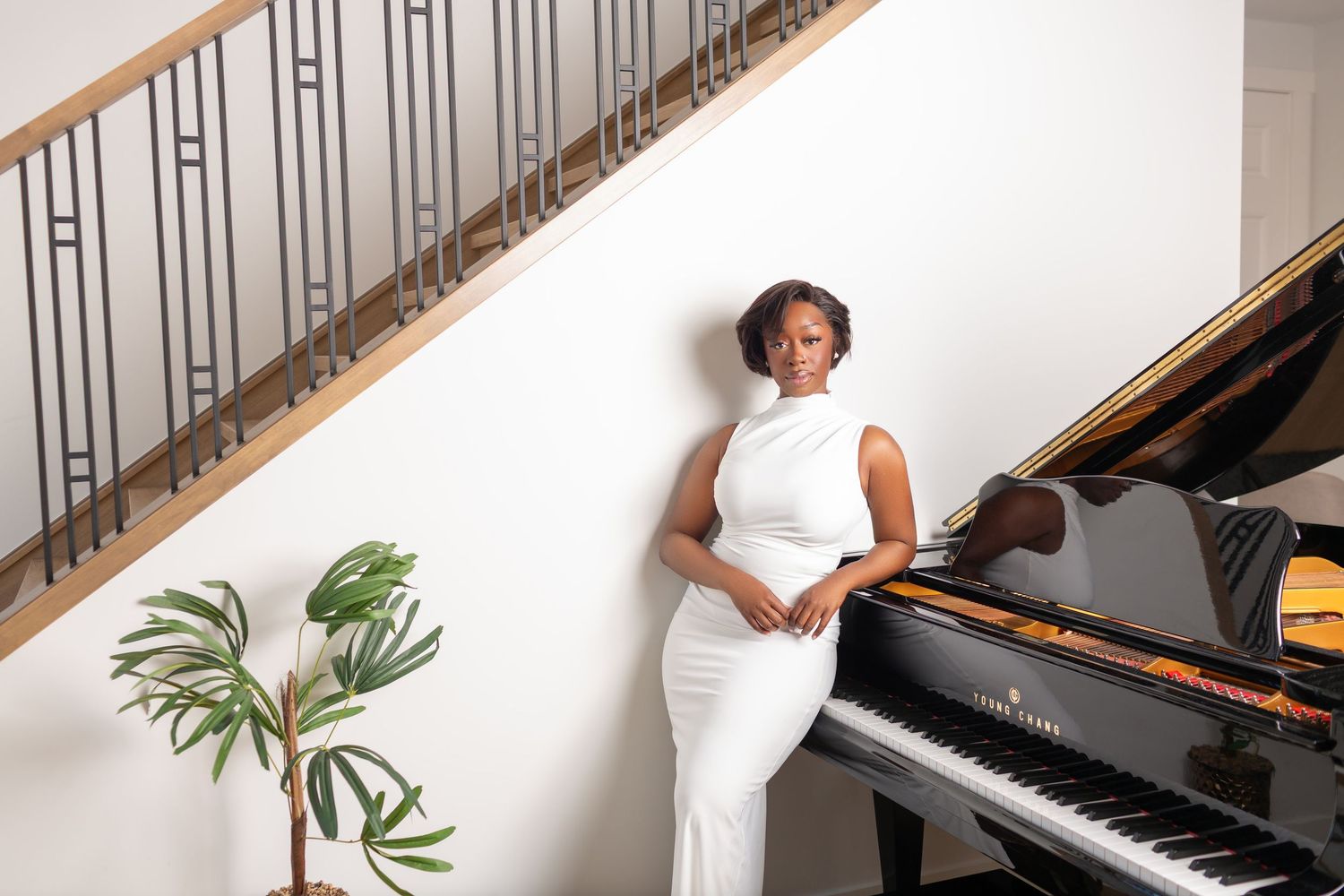 Elegant woman in white dress posing beside grand piano near staircase.