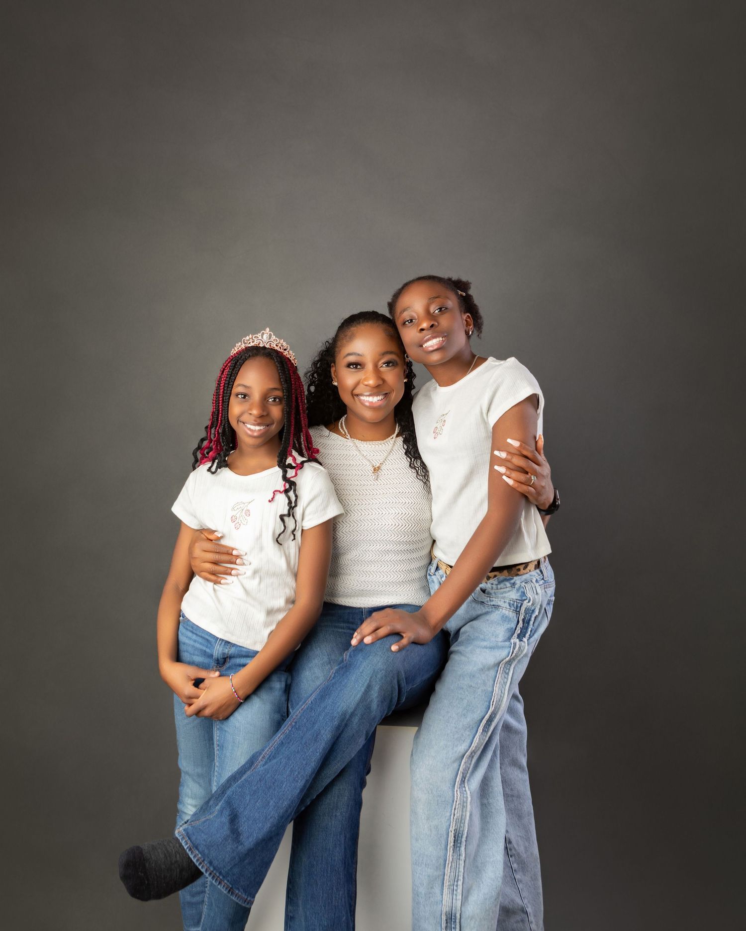 Three girls posing together on white cube in white tops and jeans in studio portrait.