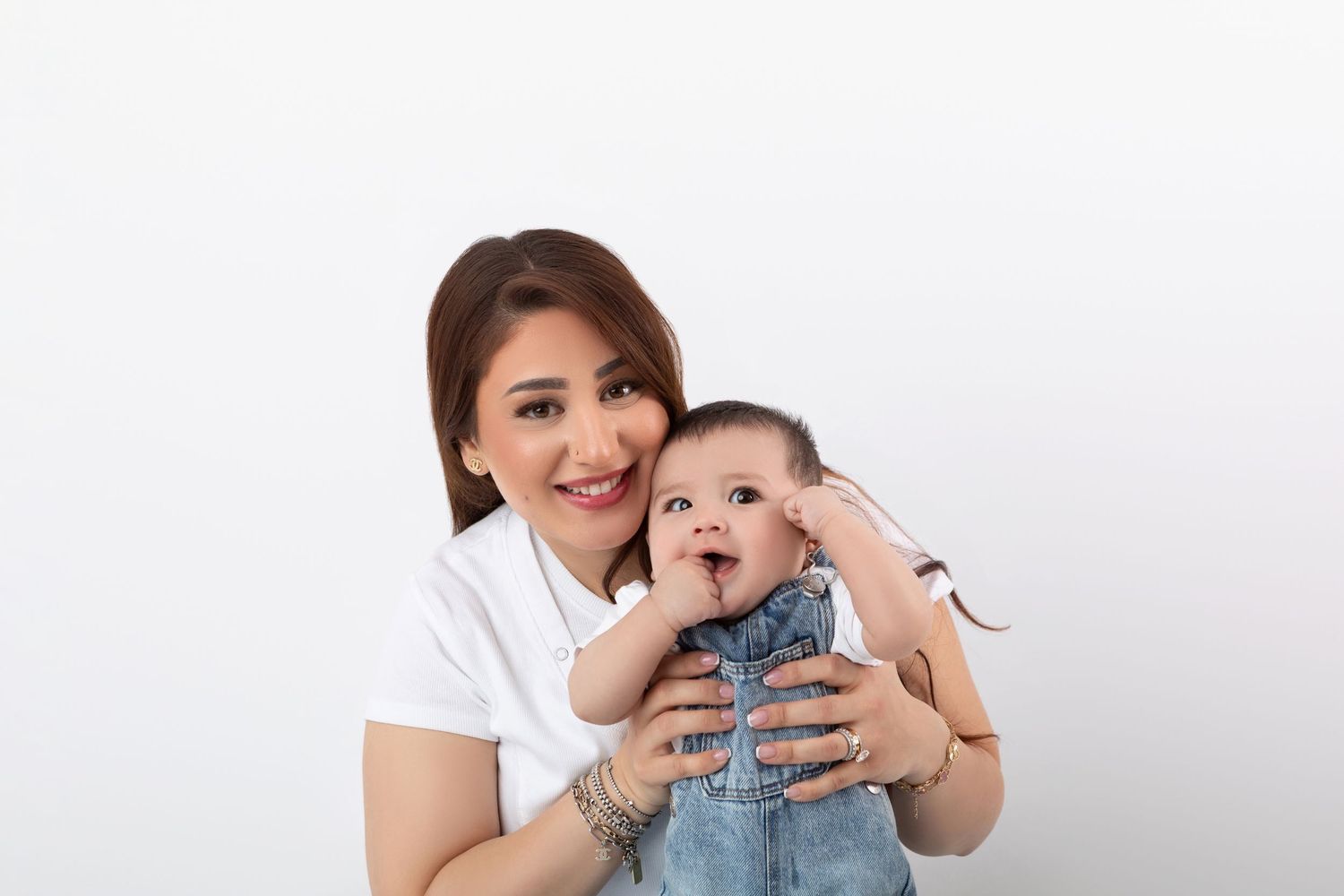 Joyful mother in white tee holds laughing baby boy in denim overalls against white backdrop.