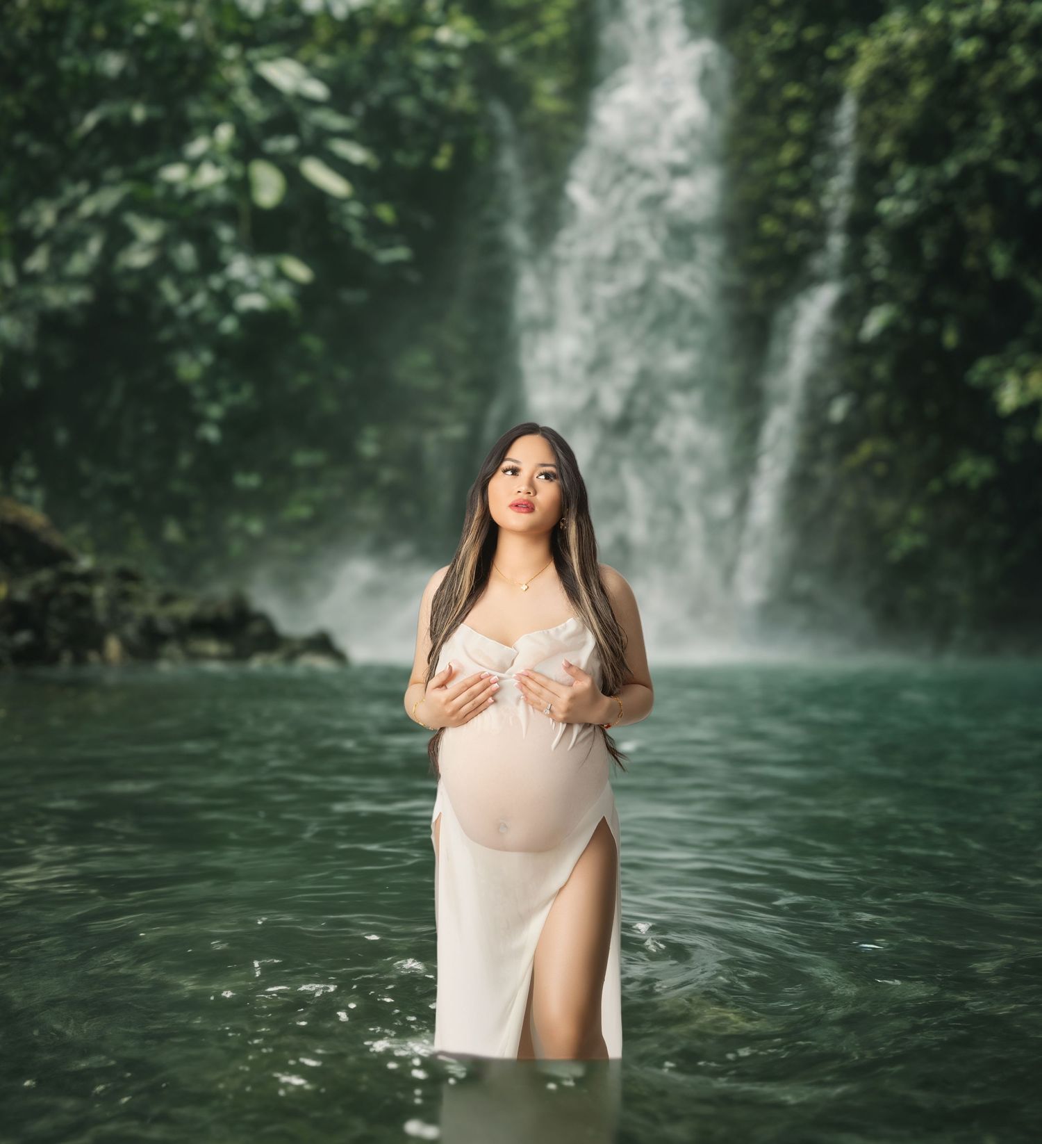 Pregnant woman in white dress standing in tropical waterfall pool surrounded by lush greenery.