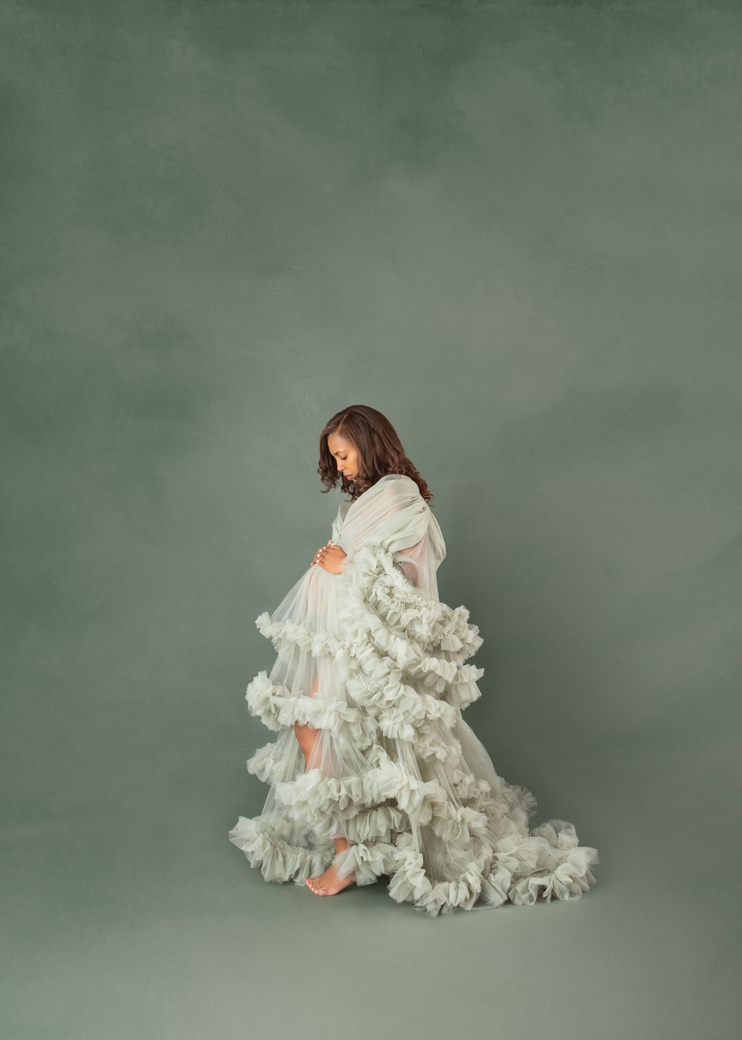 Pregnant woman in flowing white ruffled gown poses gracefully against sage green backdrop in maternity portrait.