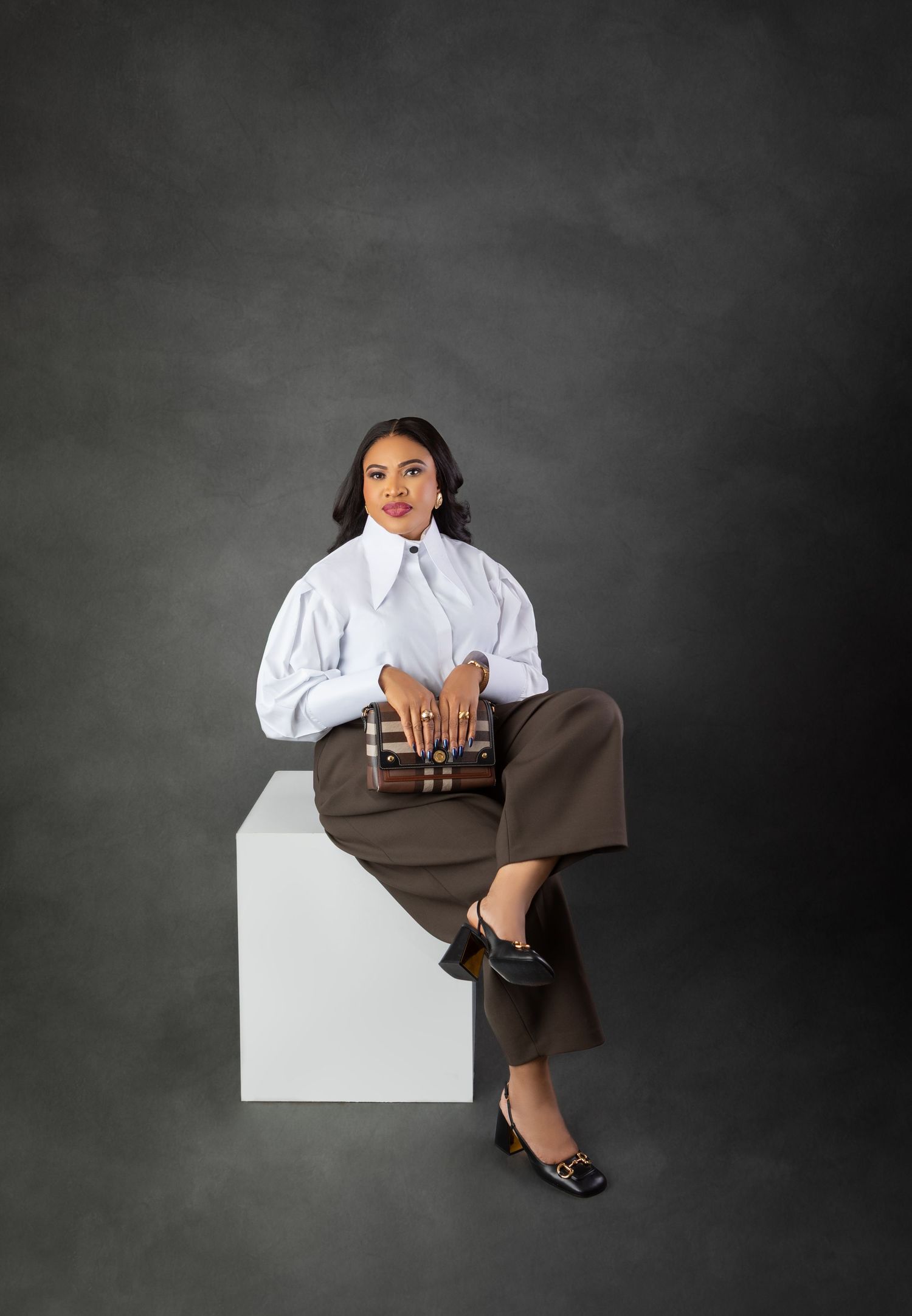 Woman in white blouse and brown trousers sits on white cube holding Burberry clutch against dark gray backdrop.