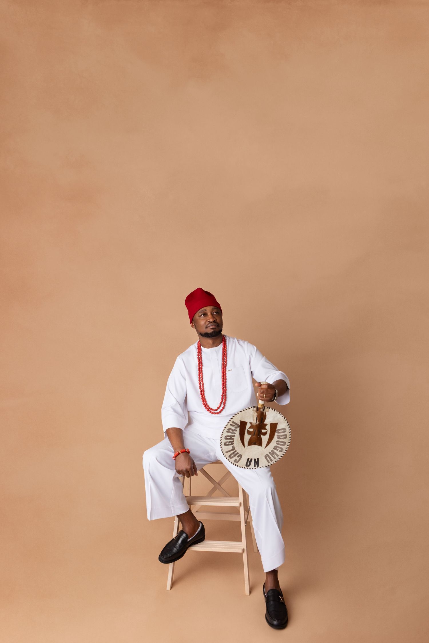 Man in white traditional Nigerian attire with red cap and beaded necklace sitting on stool holding decorative fan.