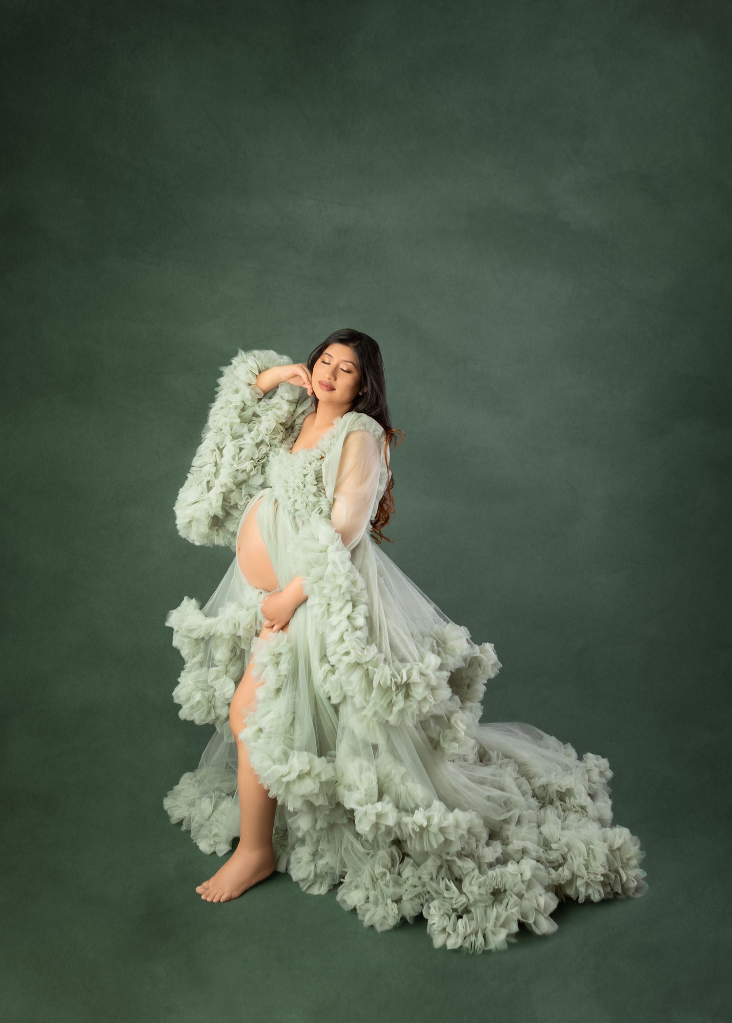 Pregnant woman in flowing mint green ruffled tulle maternity gown poses gracefully against a dark green studio backdrop.