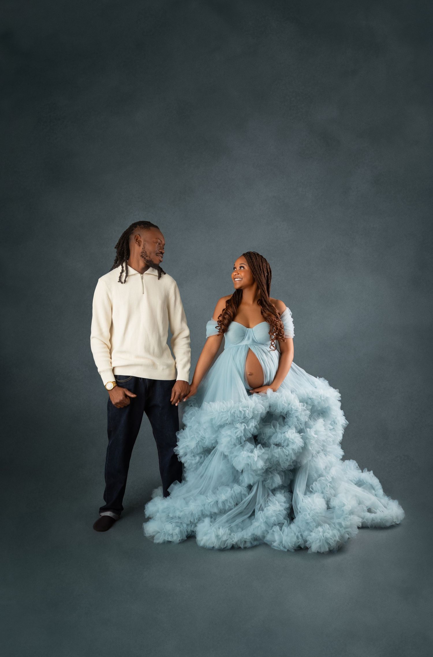 Pregnant woman in flowing light blue tulle gown holds hands with man in cream hoodie during maternity photo session.