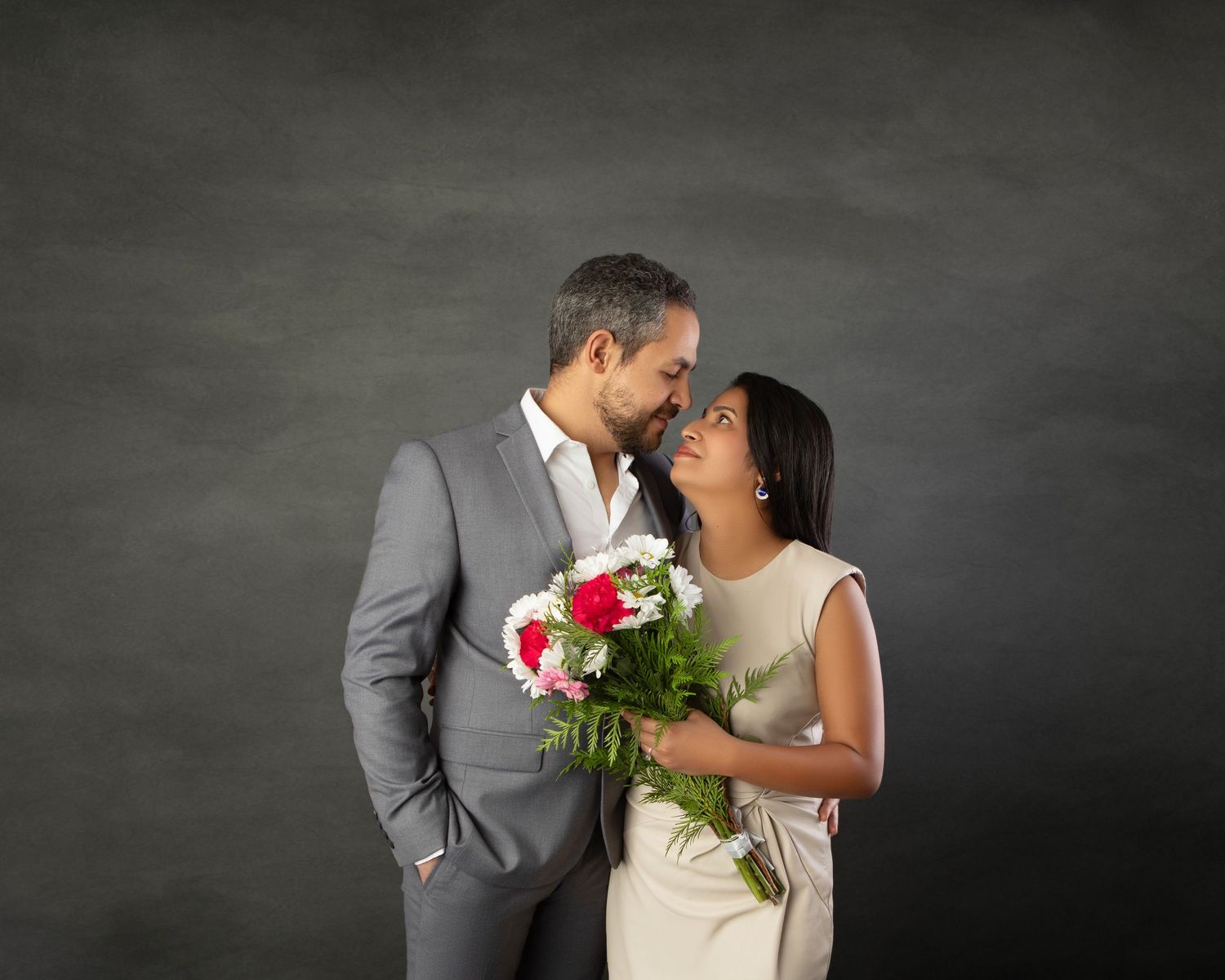 Romantic couple gazing at each other holding flowers against dark gray studio background.