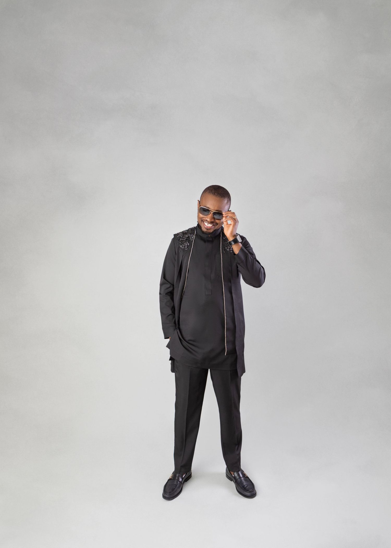 Man in all-black outfit and sunglasses smiling while talking on phone against gray background.