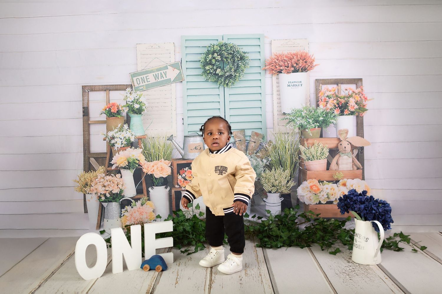 Toddler in yellow jacket stands before floral backdrop with ONE sign for first birthday photo.