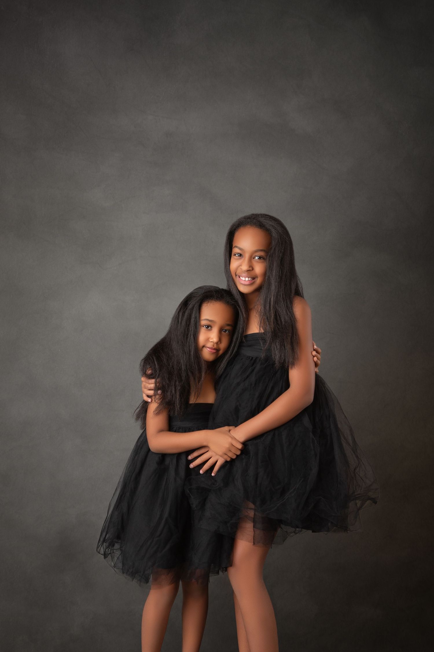 Two young girls in matching black tulle dresses embrace warmly against a gray studio backdrop.