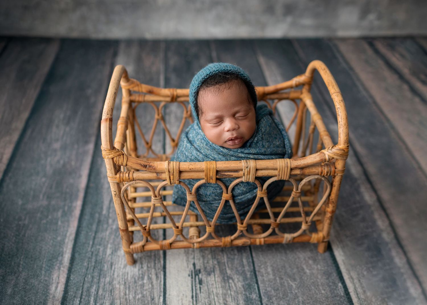 Newborn baby wrapped in teal swaddle sleeping in a small wicker basket on wood floor.