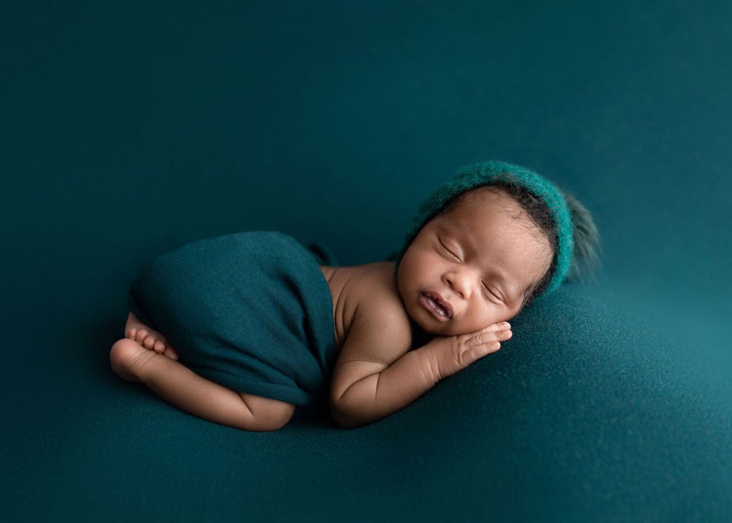 Sleeping newborn baby in teal wrap and knit hat on teal background.