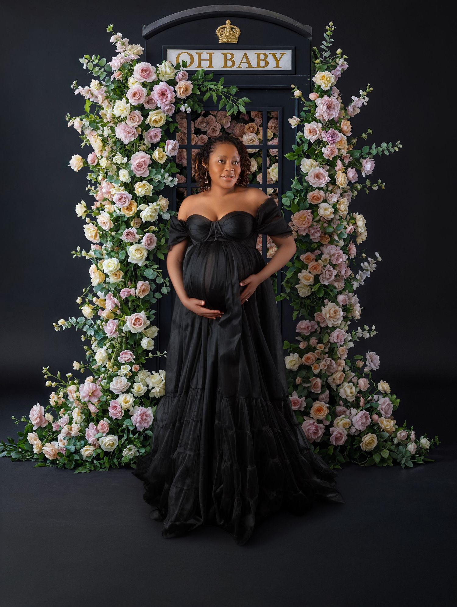 Pregnant woman in black off-shoulder gown stands before floral arch with Oh Baby sign.