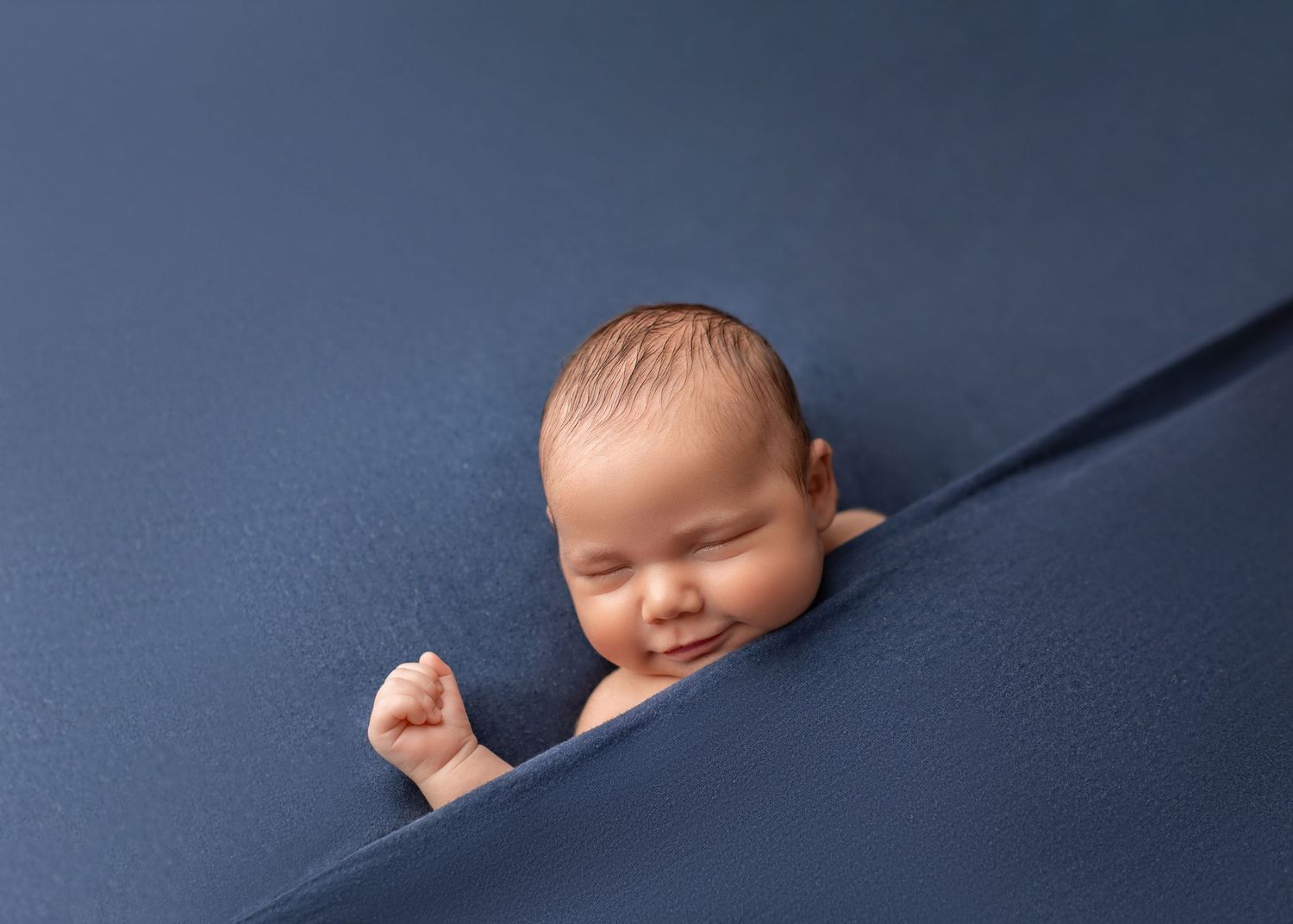 Smiling newborn baby boy peeking out from navy blue fabric wrap.
