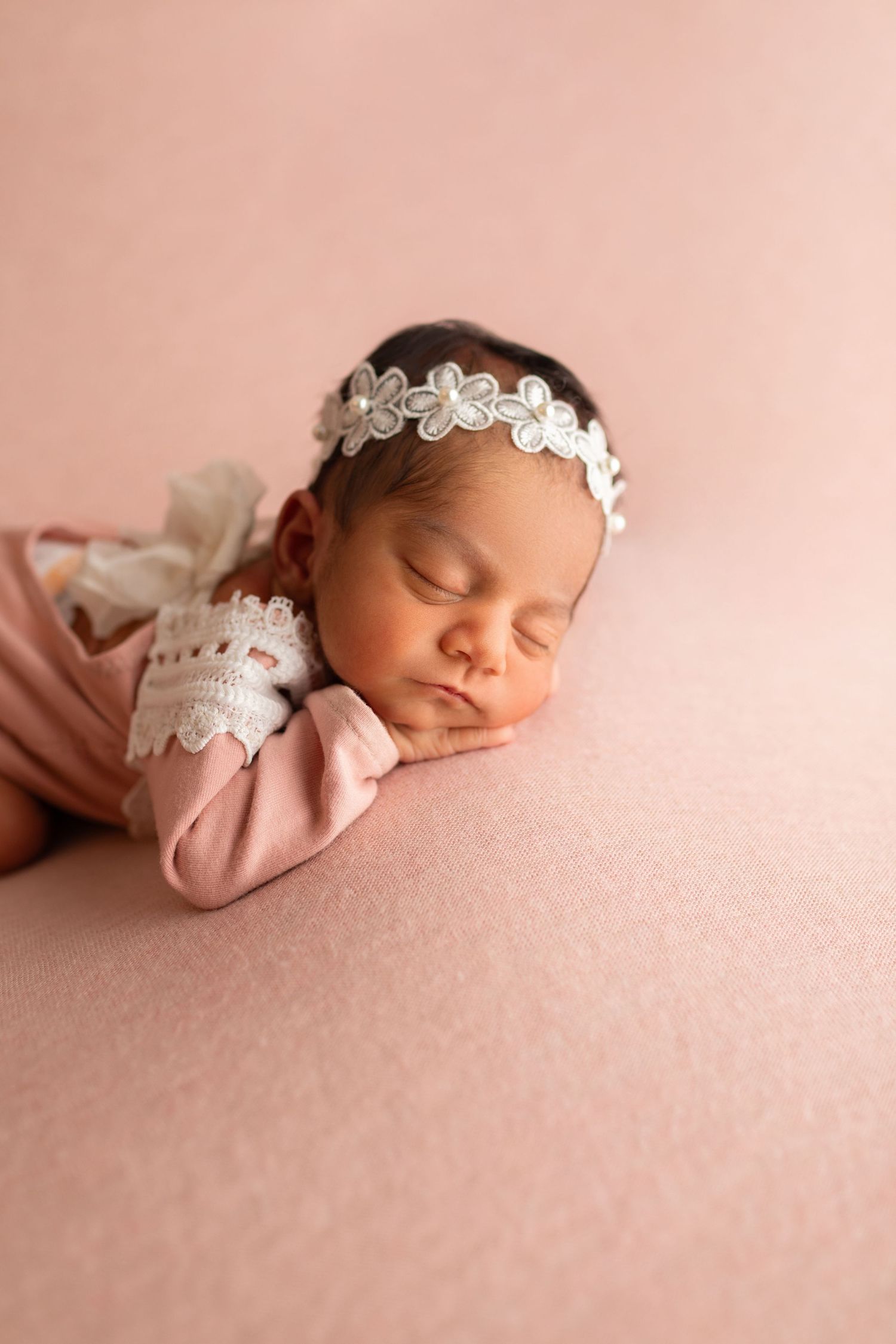 Newborn baby girl sleeping on pink backdrop wearing pink outfit with lace cuffs and white floral headband.
